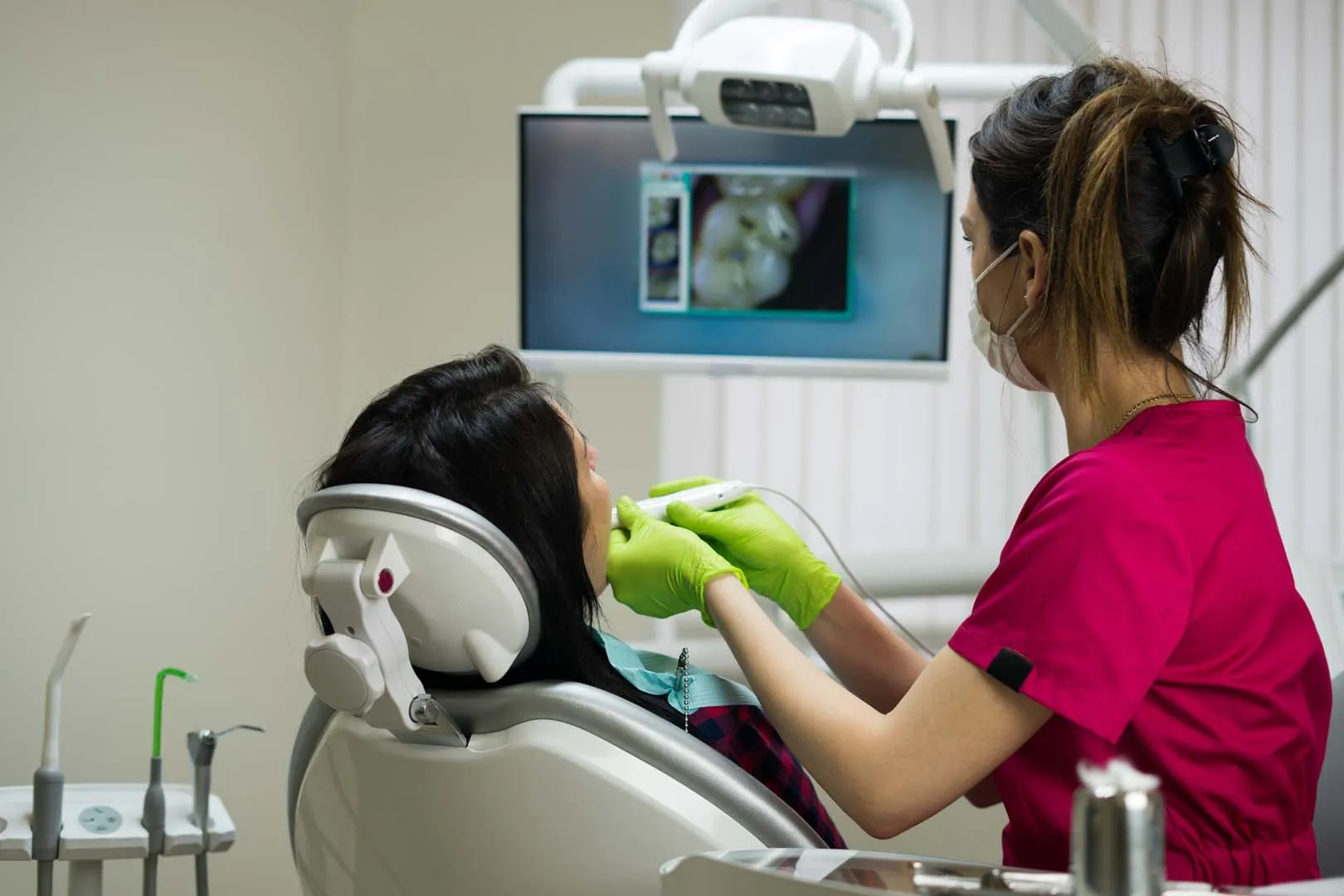 Dentist wearing pink scrubs and green gloves examining patient's teeth using an intraoral camera.