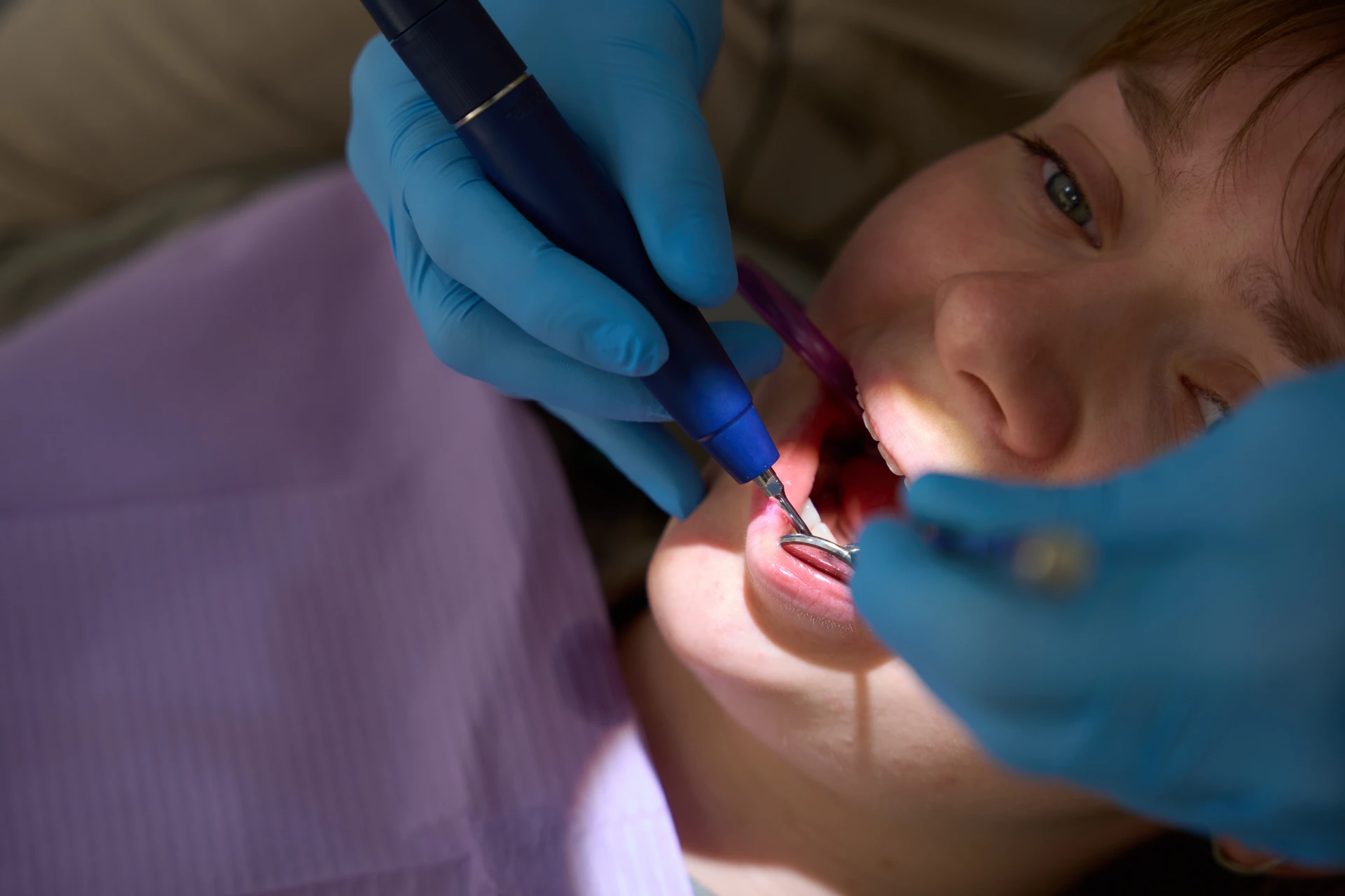 Dentist wearing blue gloves using dental tools inside a young woman's open mouth.