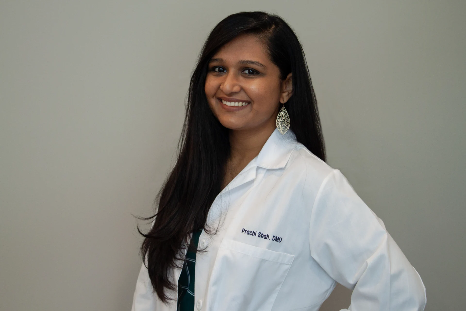 Smiling woman with long dark hair wearing a white coat with name and title embroidery, standing