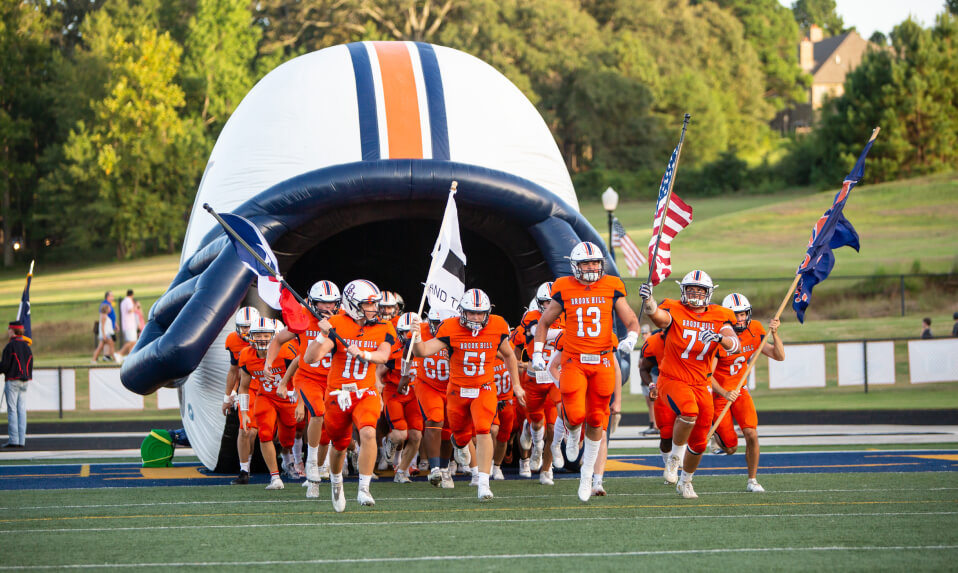 The Brook Hill School football team running out onto the field through a large inflatable helmet.