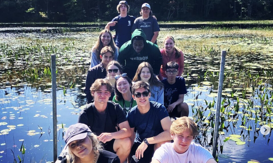 Hebron Academy students posing for a picture on a dock surrounded by lily pads on the water.