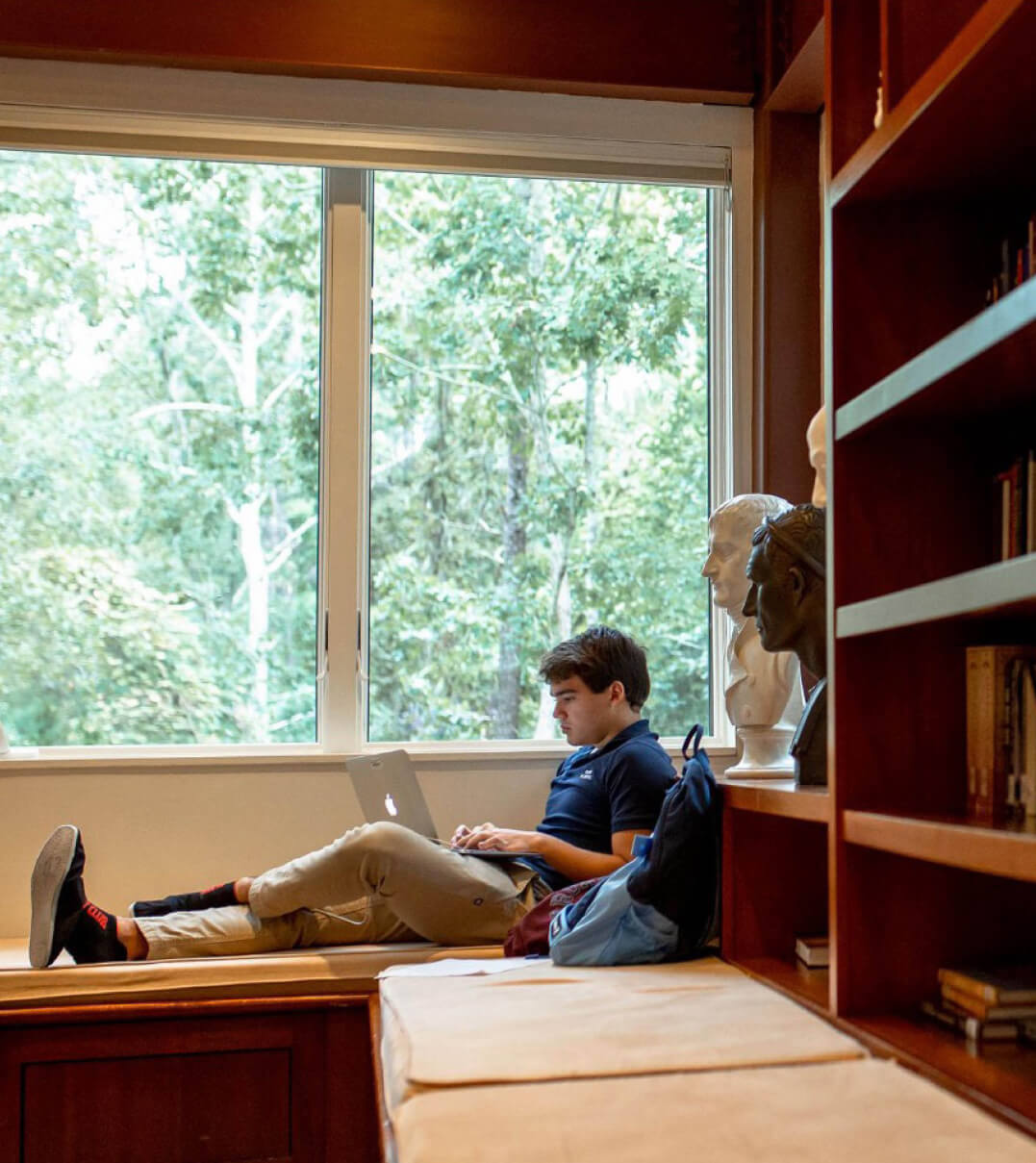 A Ross School student working on his laptop while seated next to a window.