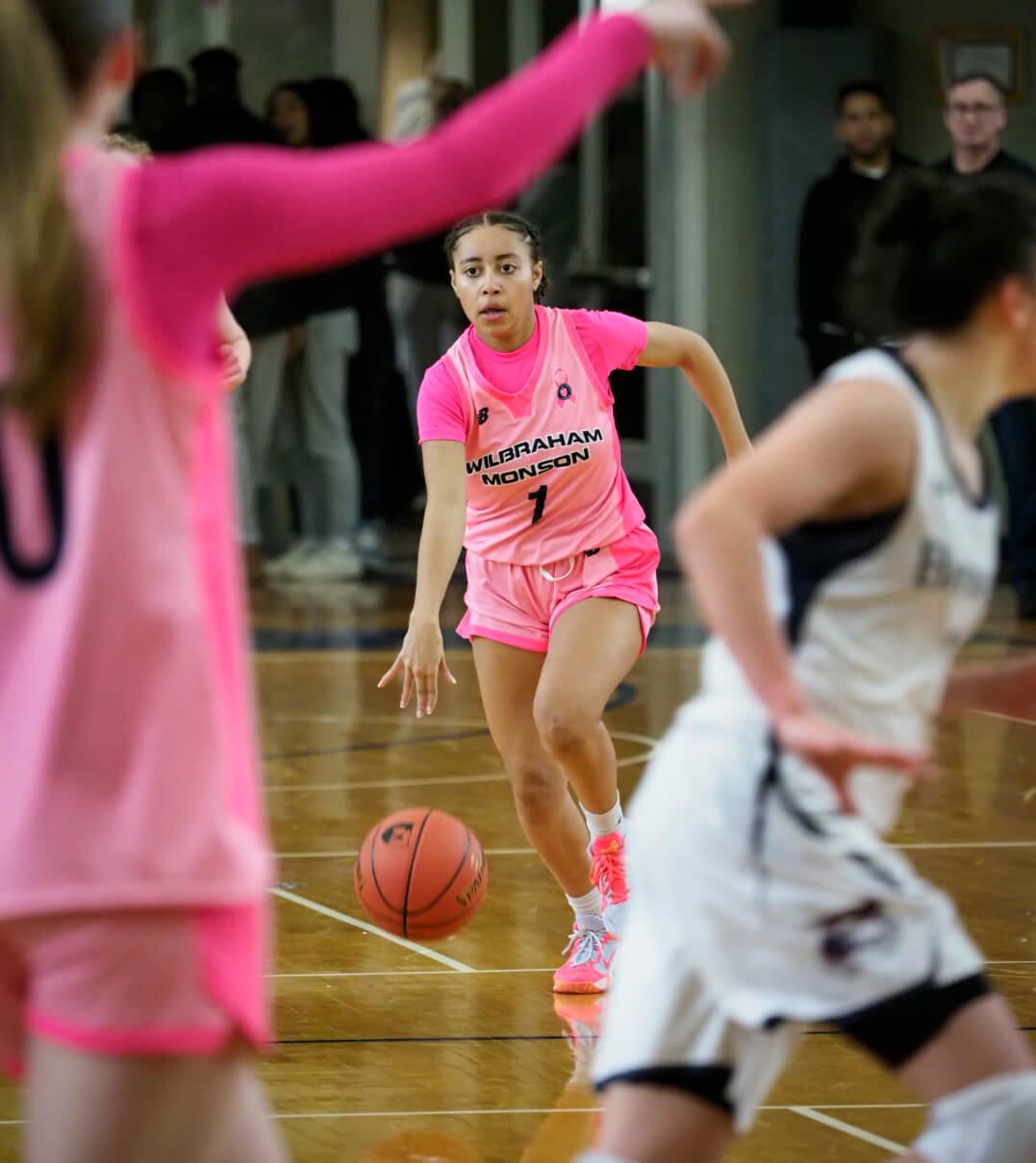 A WMA woman's basketball player, wearing a pink breast cancer awareness jersey, dribbles down the court.