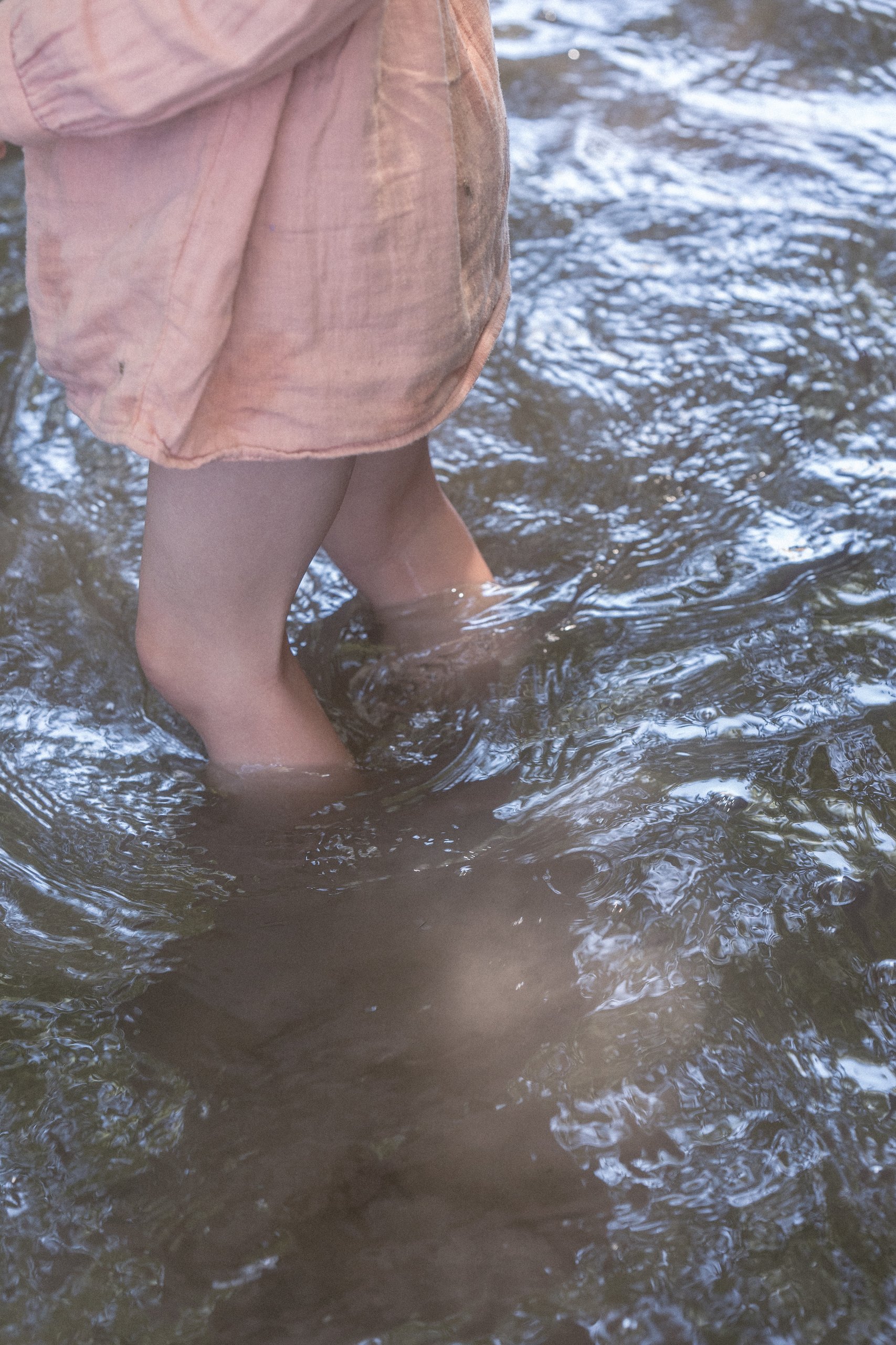 A child in a pink dress wading barefoot through a sparkling creek