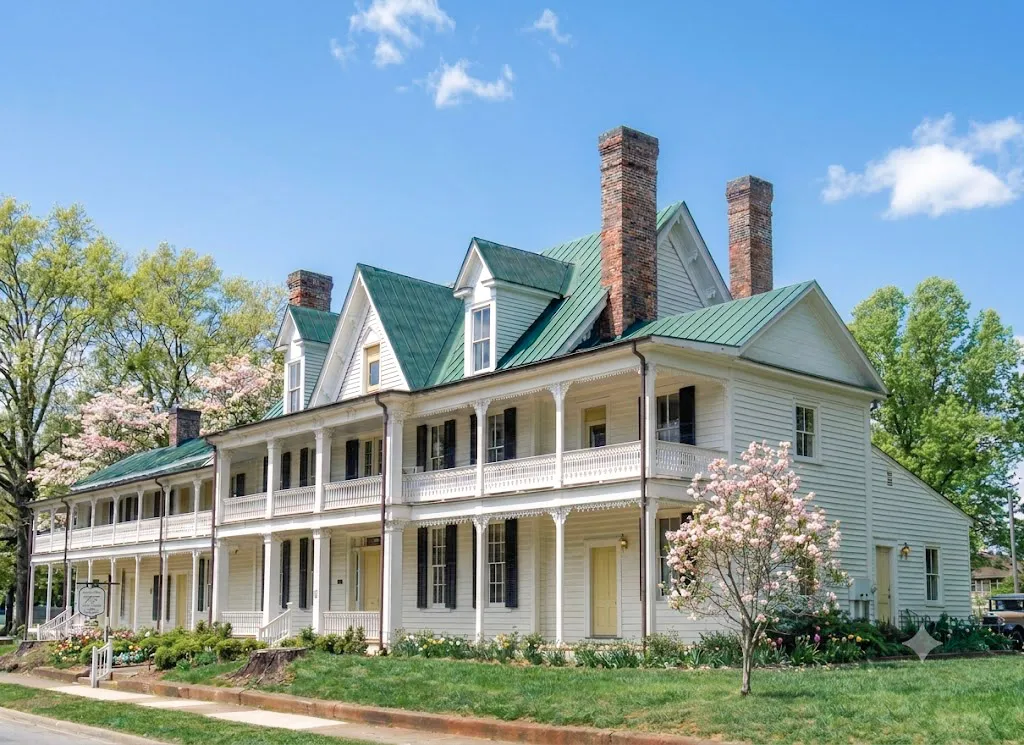 Large historic white two-story house with green metal roofs, brick chimneys, and flowering trees and shrubs.
