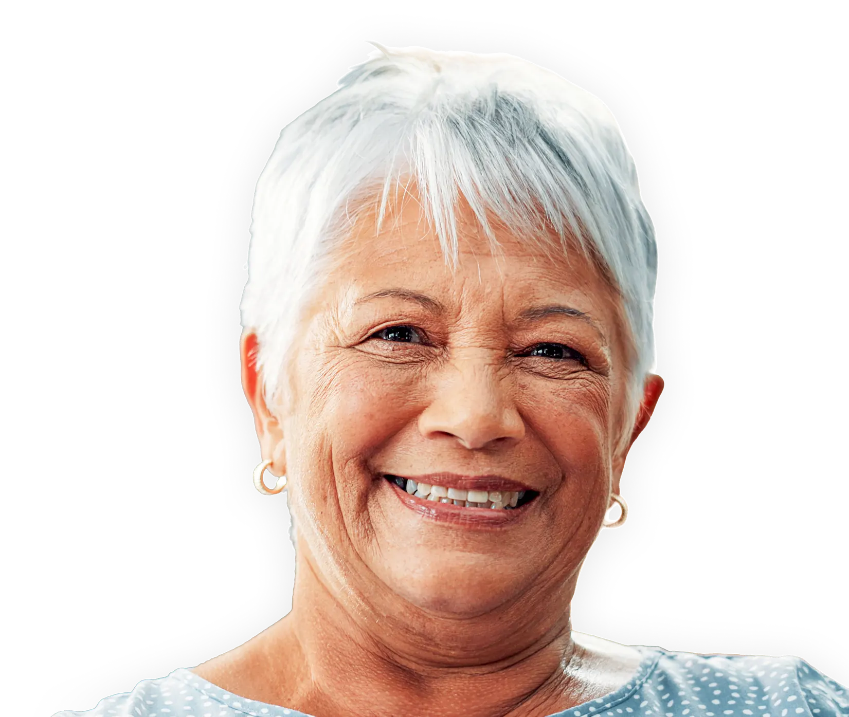 Smiling elderly woman with short white hair wearing a light blue polka dot shirt and gold hoop earrings.