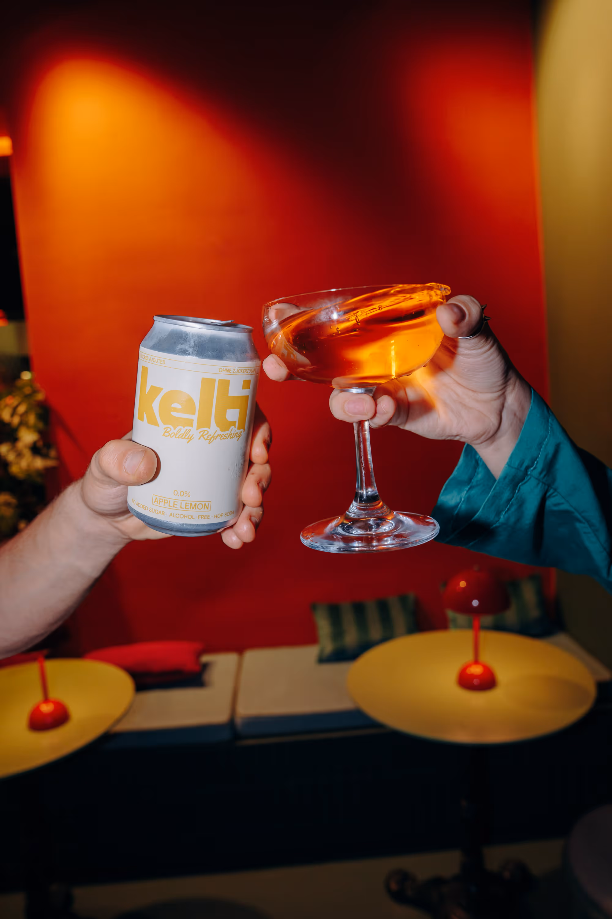 Two hands clinking a can of kelti apple lemon soda and a glass with an orange drink inside.