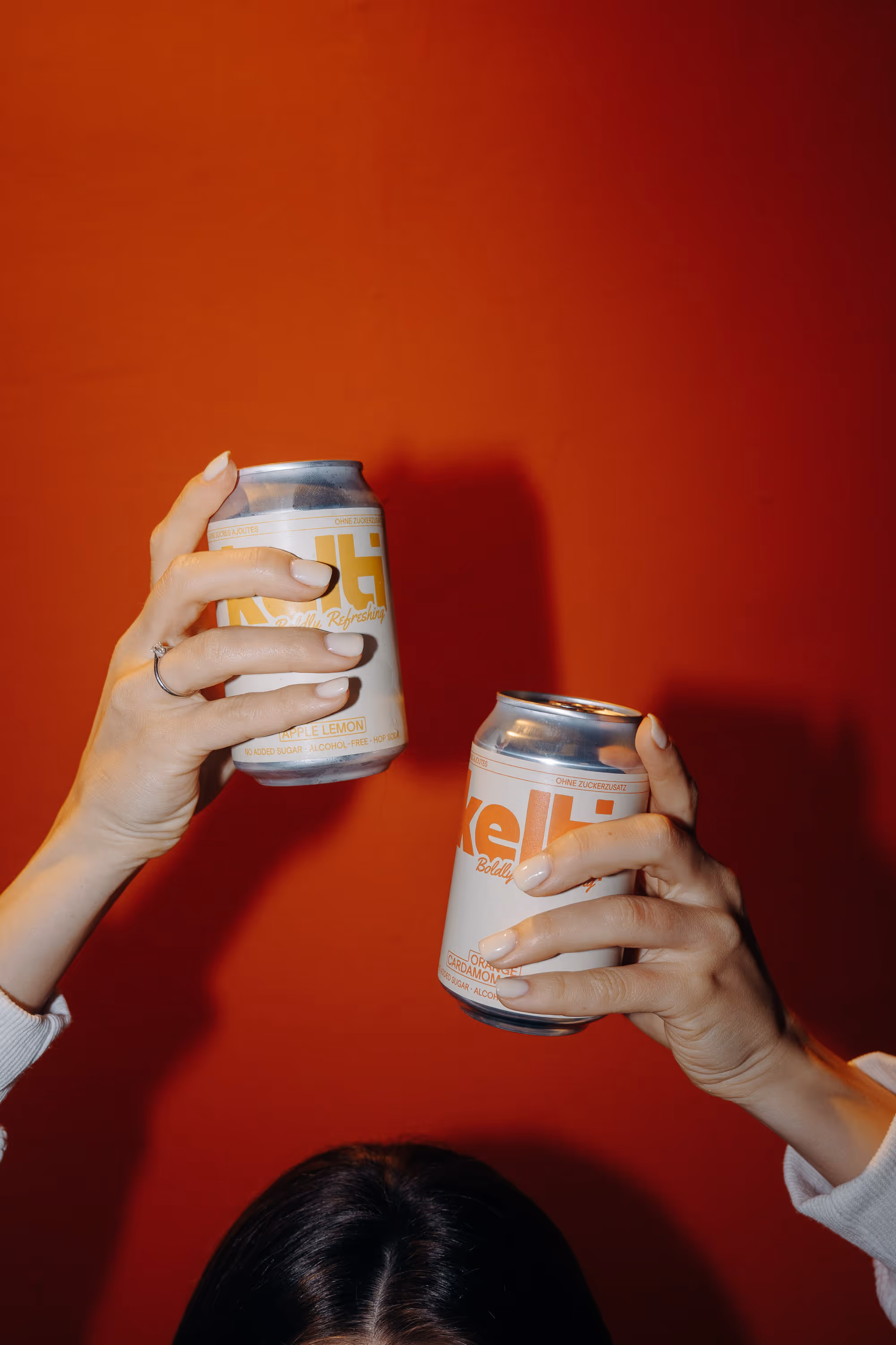 Two hands holding white Kelti beverage cans against a red background over a person’s head.