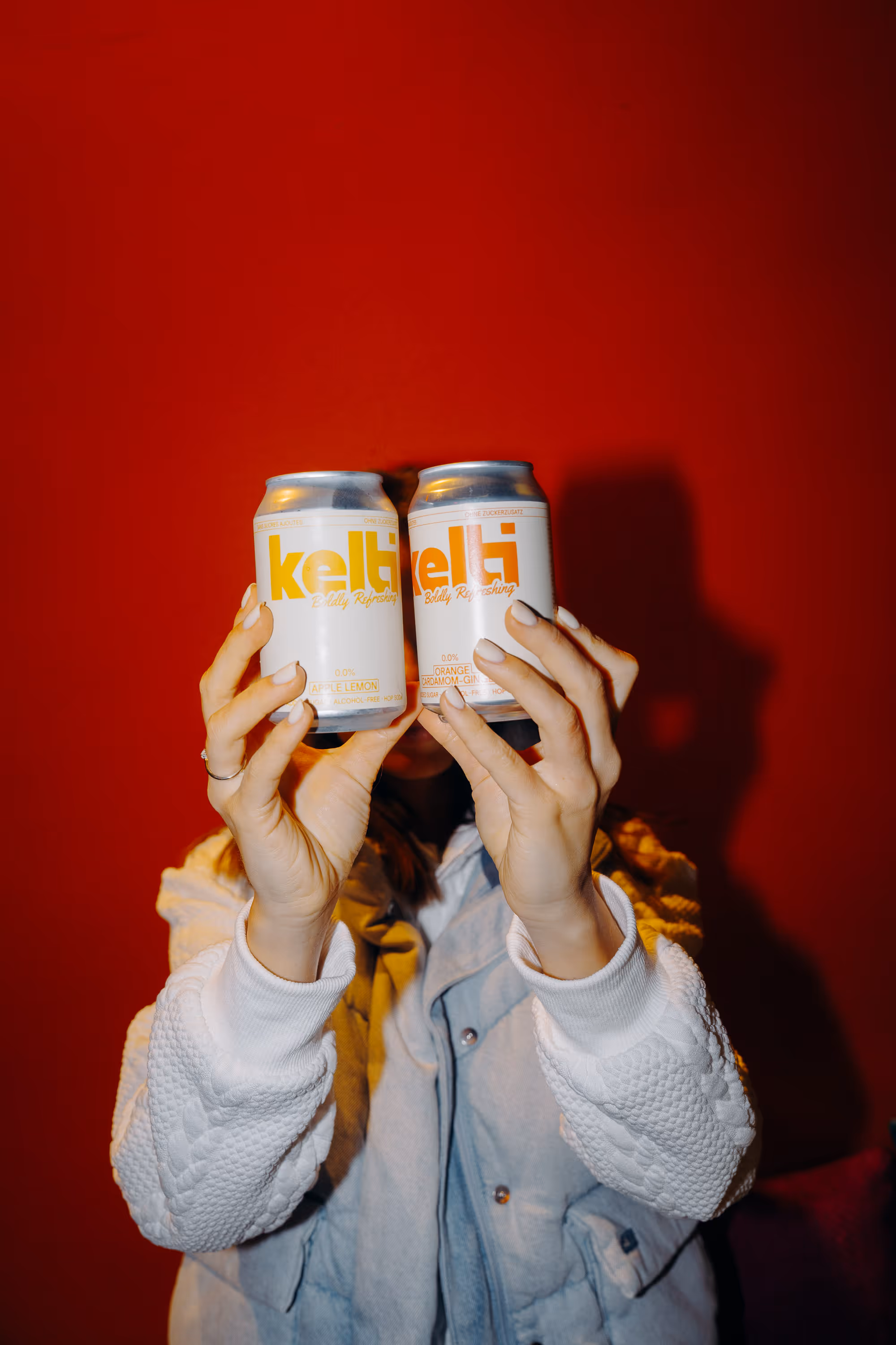 Person holding two white Kelti drink cans labeled Apple Lemon and Orange Cardamom Gin against a red background.