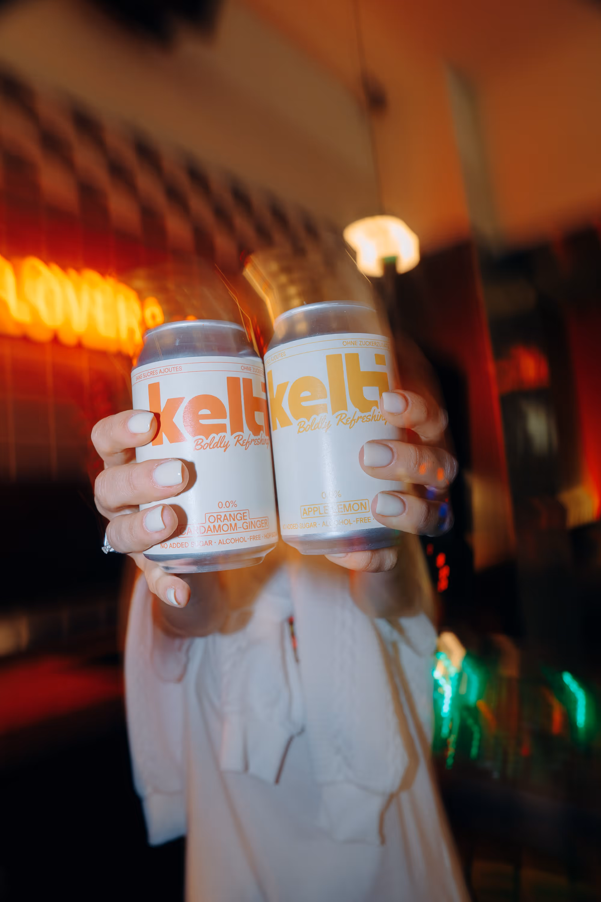 Person holding two Kelti branded cans: Orange Cardamom Ginger and Apple Lemon flavors.