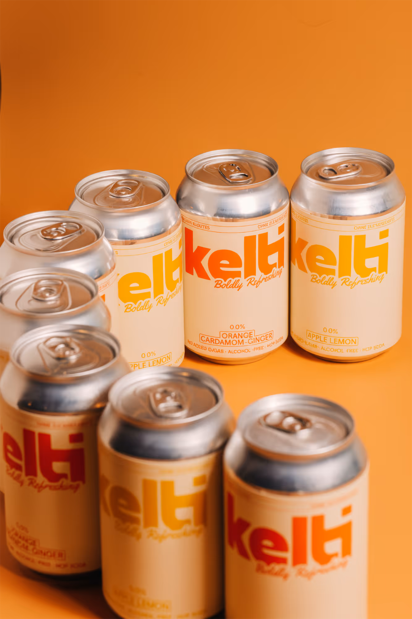 Cans of Kelti non-alcoholic soda in orange cardamom-ginger and apple lemon flavors on orange background.