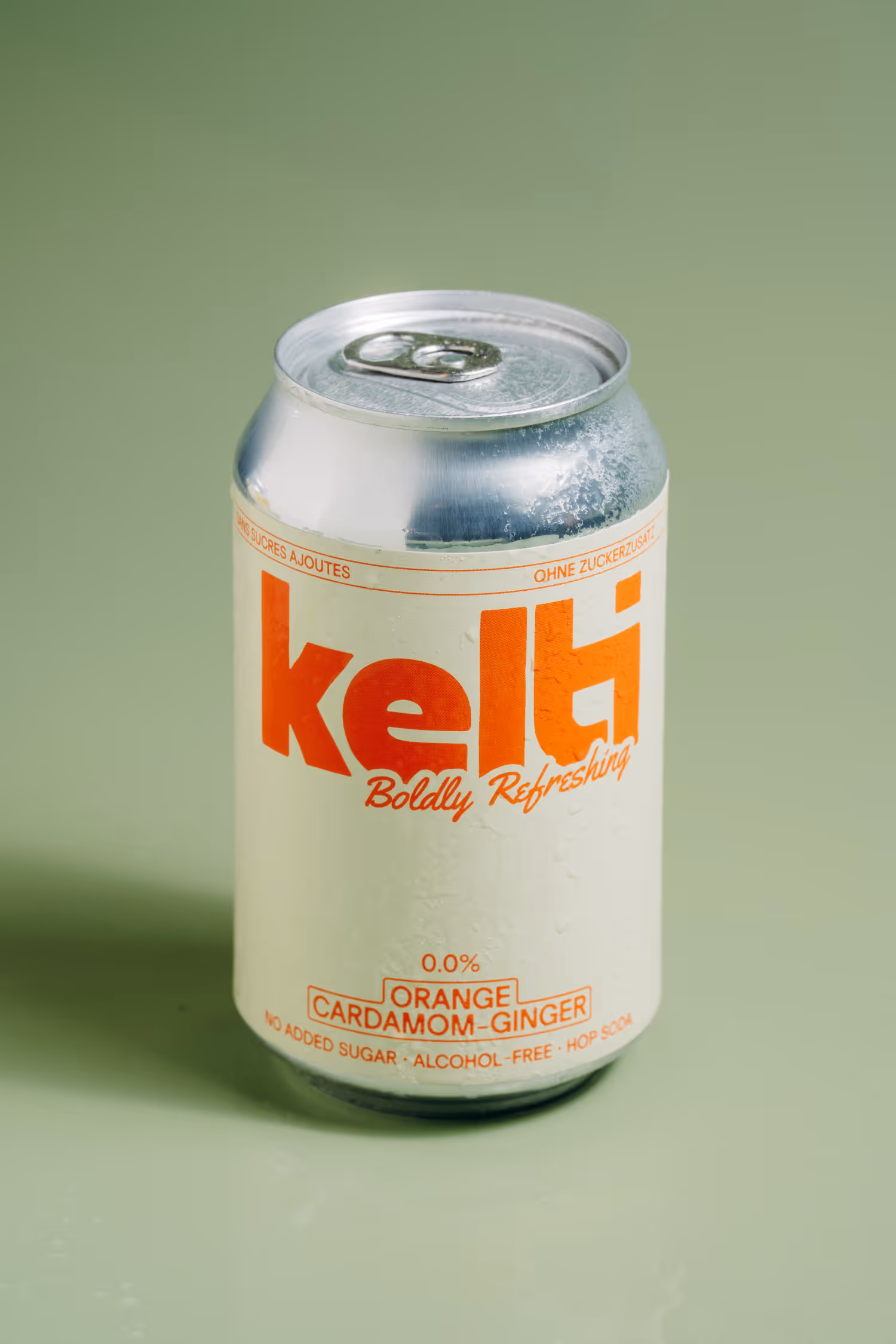 Can of Kelti orange cardamom-ginger hop soda with no added sugar, alcohol-free on green background.