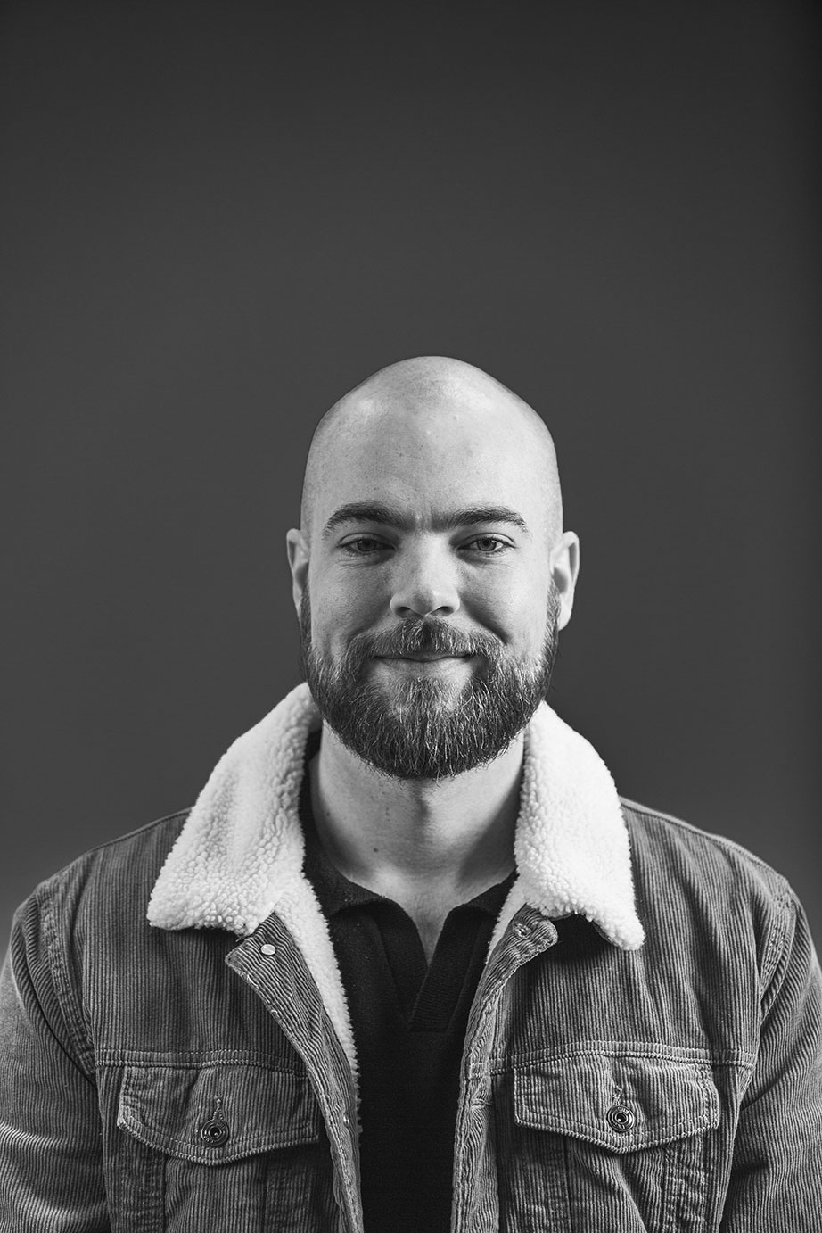 Smiling bald man with beard wearing a jacket with a fluffy collar against a plain background.