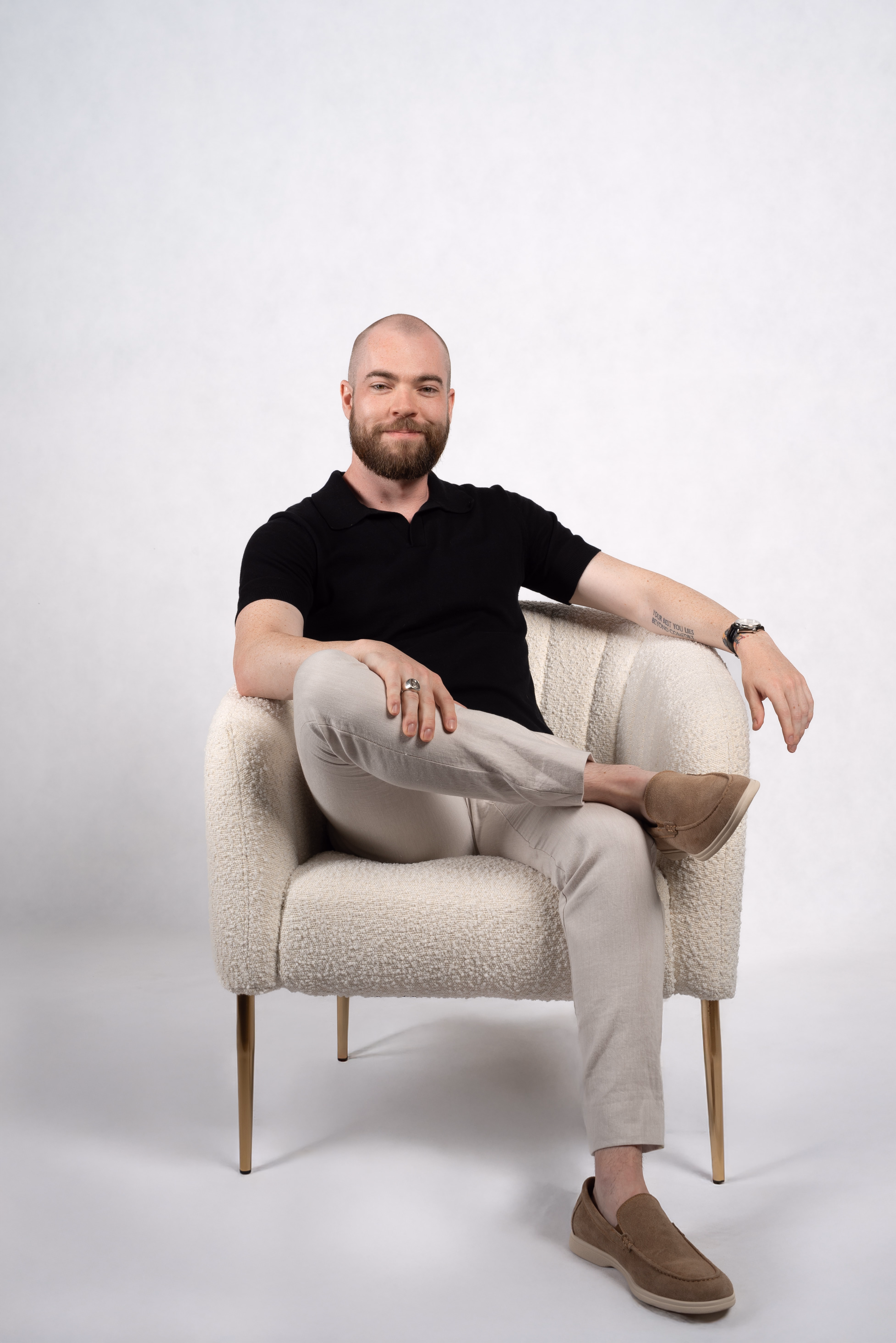 Bearded man in black shirt, beige pants, and brown shoes sits cross-legged in a textured white armchair.