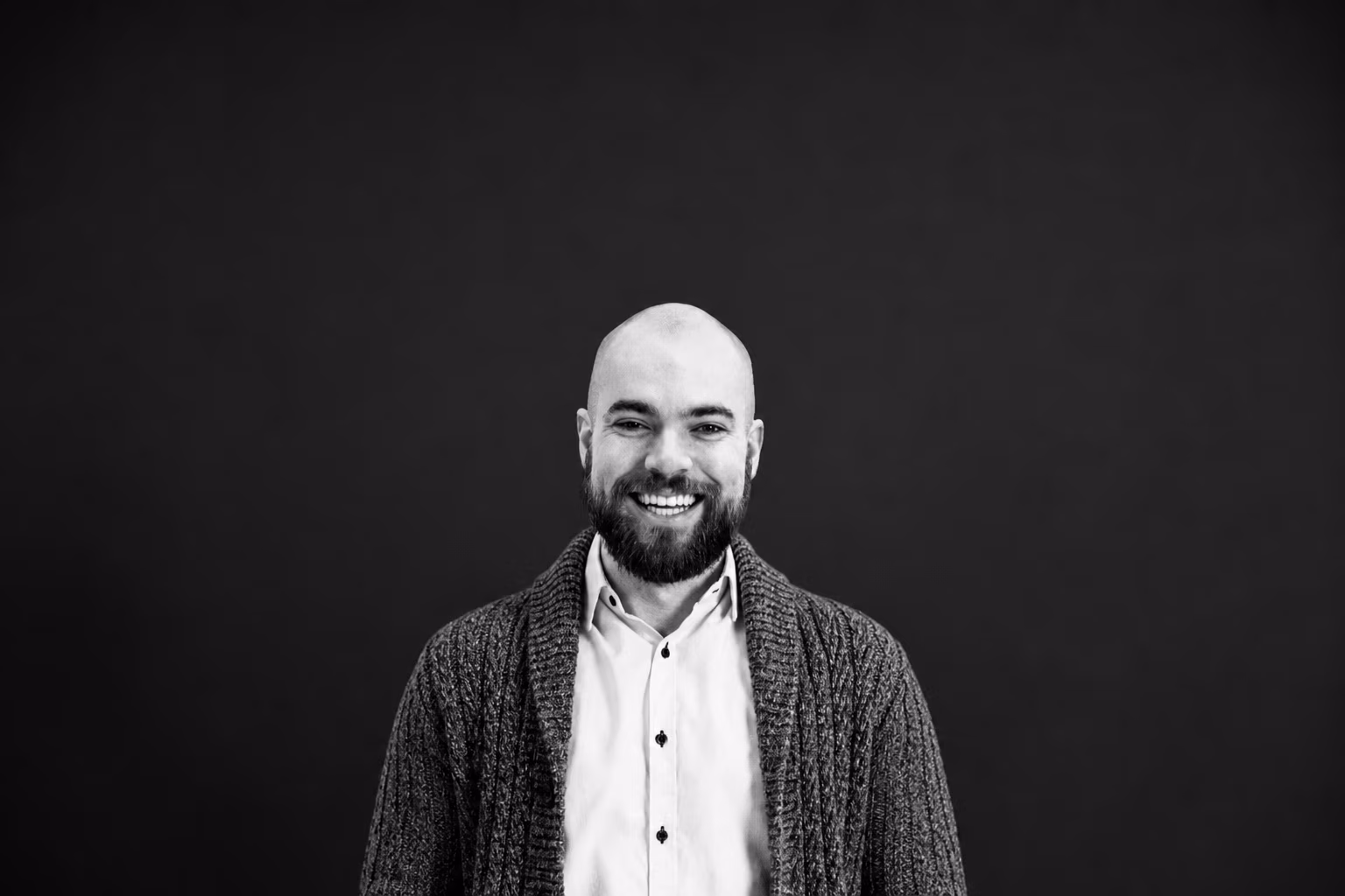 Smiling bald man with beard wearing a button-up shirt and knit cardigan against dark background.