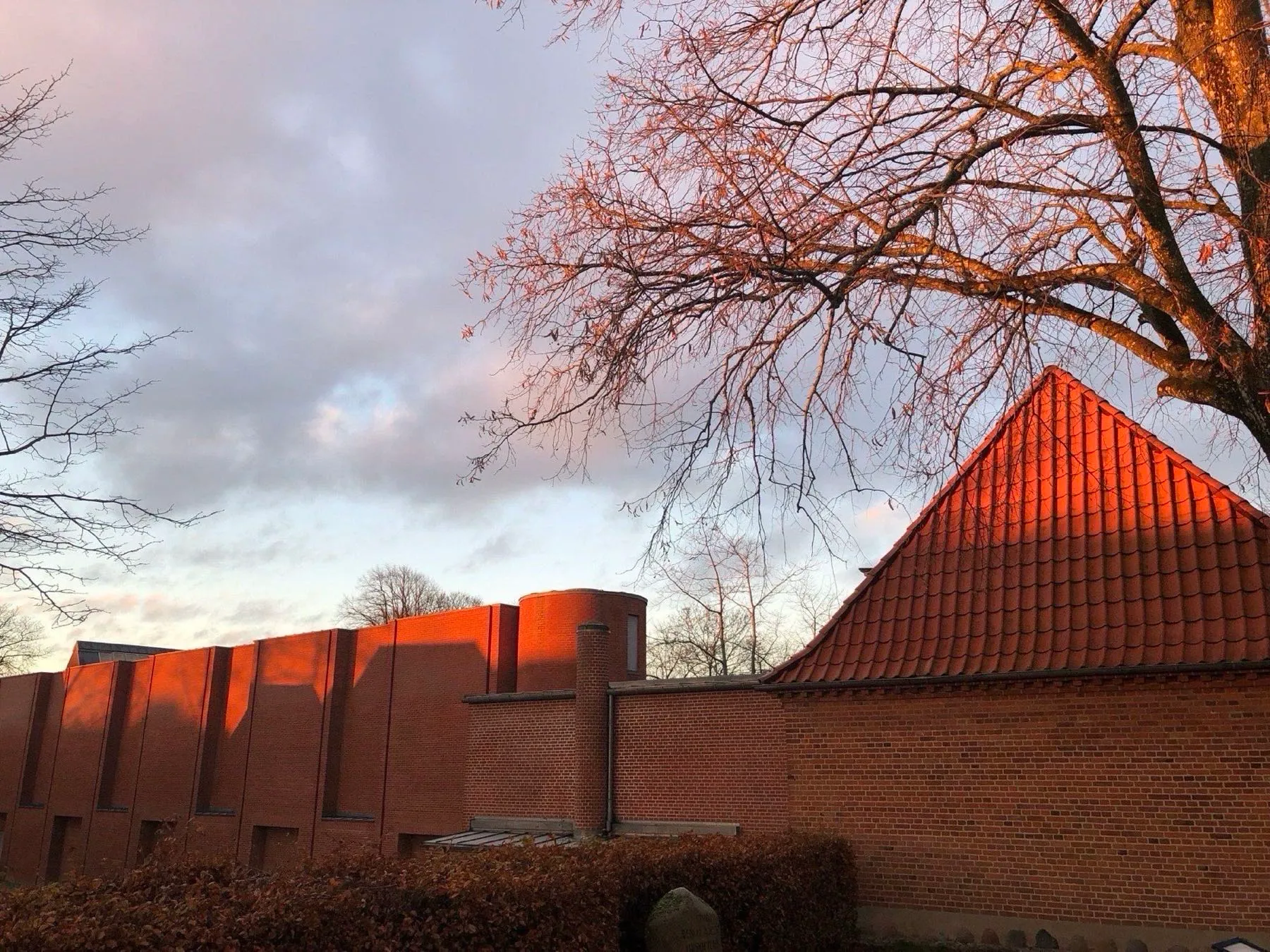 Museets murstensfacade og røde tegltag i lav aftensol med et bladløst træ mod skyet himmel