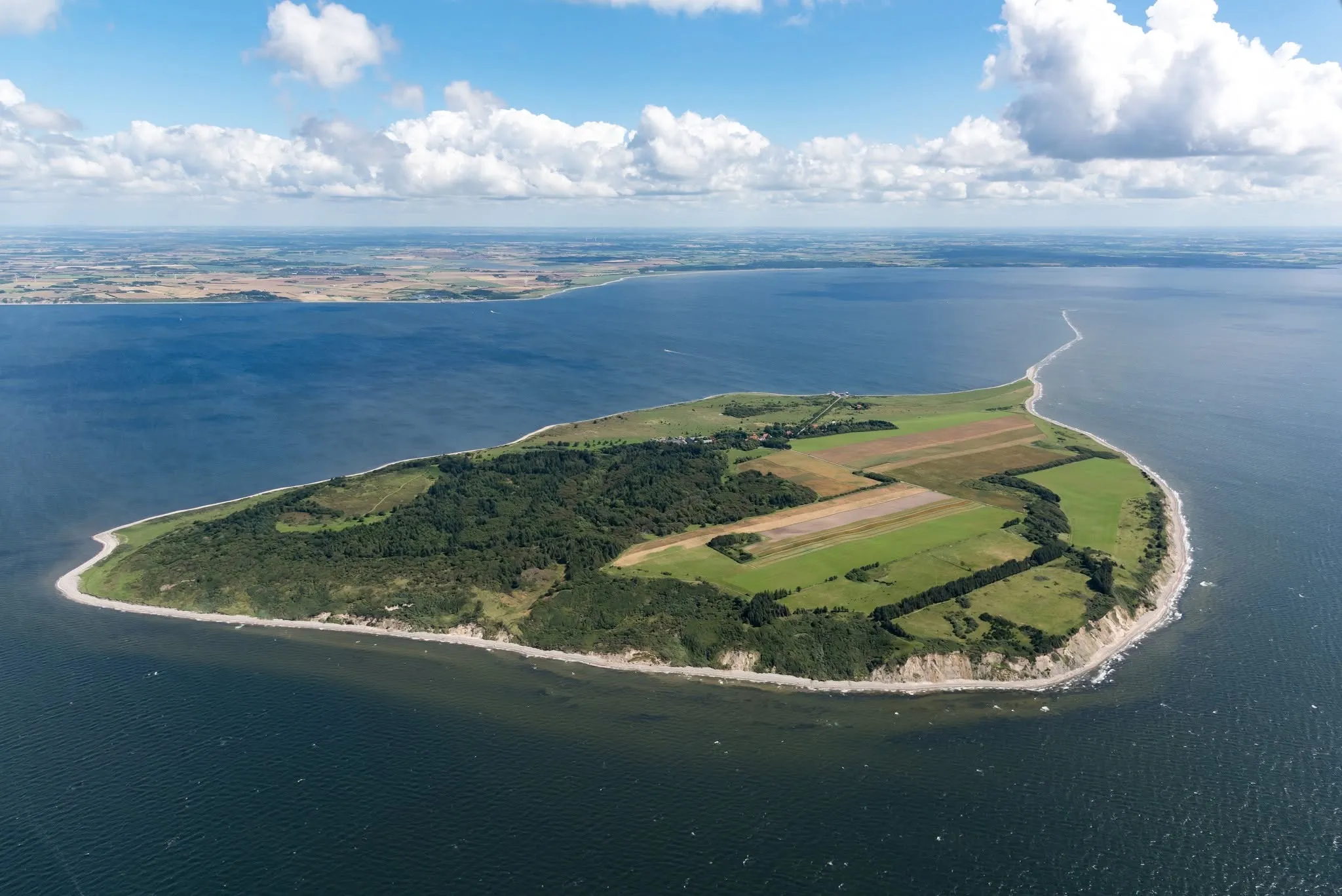 Luftfoto af en grøn ø med marker, skov og lys strandkant omgivet af blåt hav under skyer