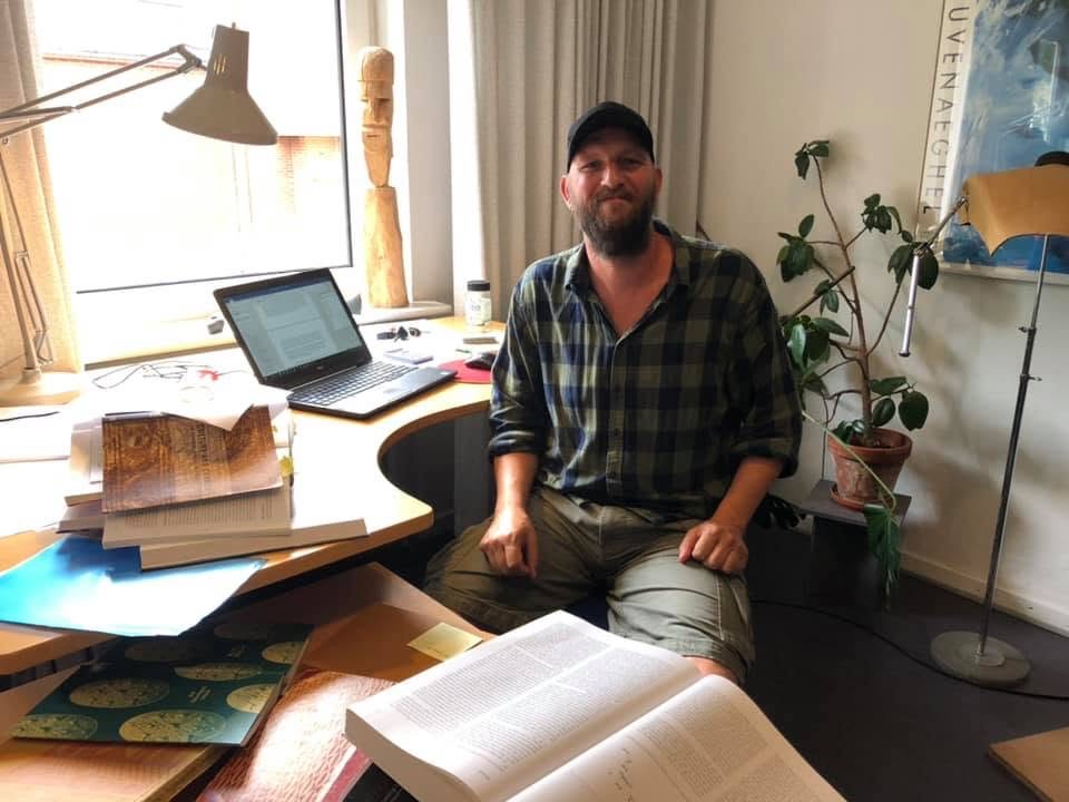 Mand med kasket sidder ved et skrivebord fyldt med bøger, papirer og en bærbar computer