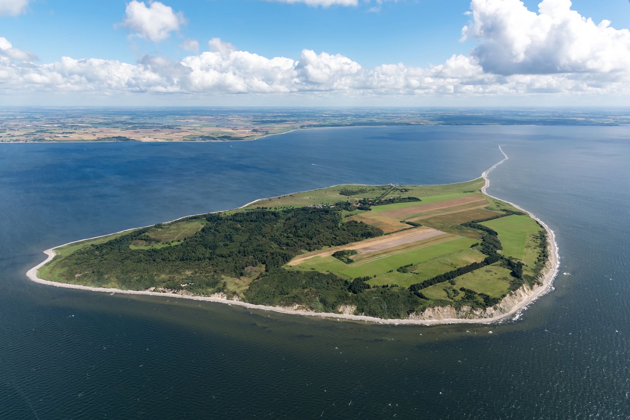 Luftfoto af en grøn ø med marker, skov og lys strandkant omgivet af blåt hav under skyer