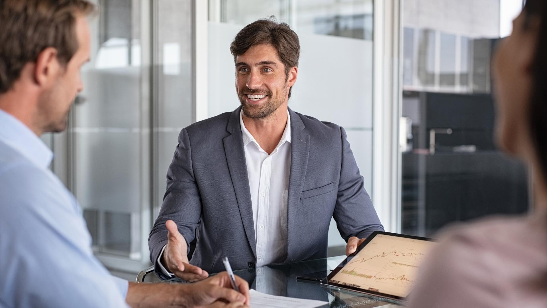 Smiling businessman shows financial charts on tablet during meeting with colleagues at office.