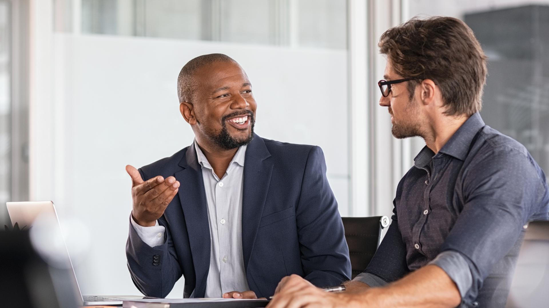 Two men in office discussing, one smiling and gesturing while the other listens attentively.