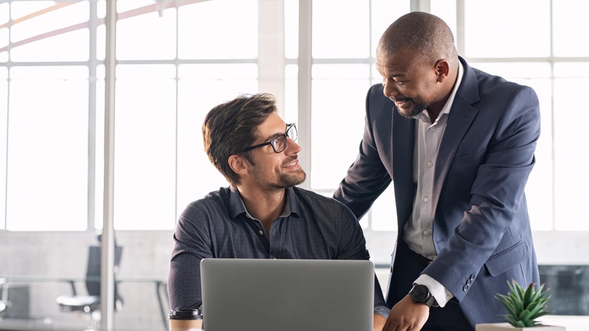 Man in glasses sitting with laptop smiles at standing man in suit in modern office setting.