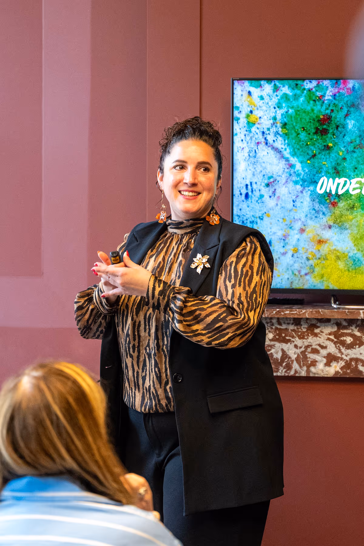 Woman with patterned blouse and black vest giving presentation in front of a colorful screen.