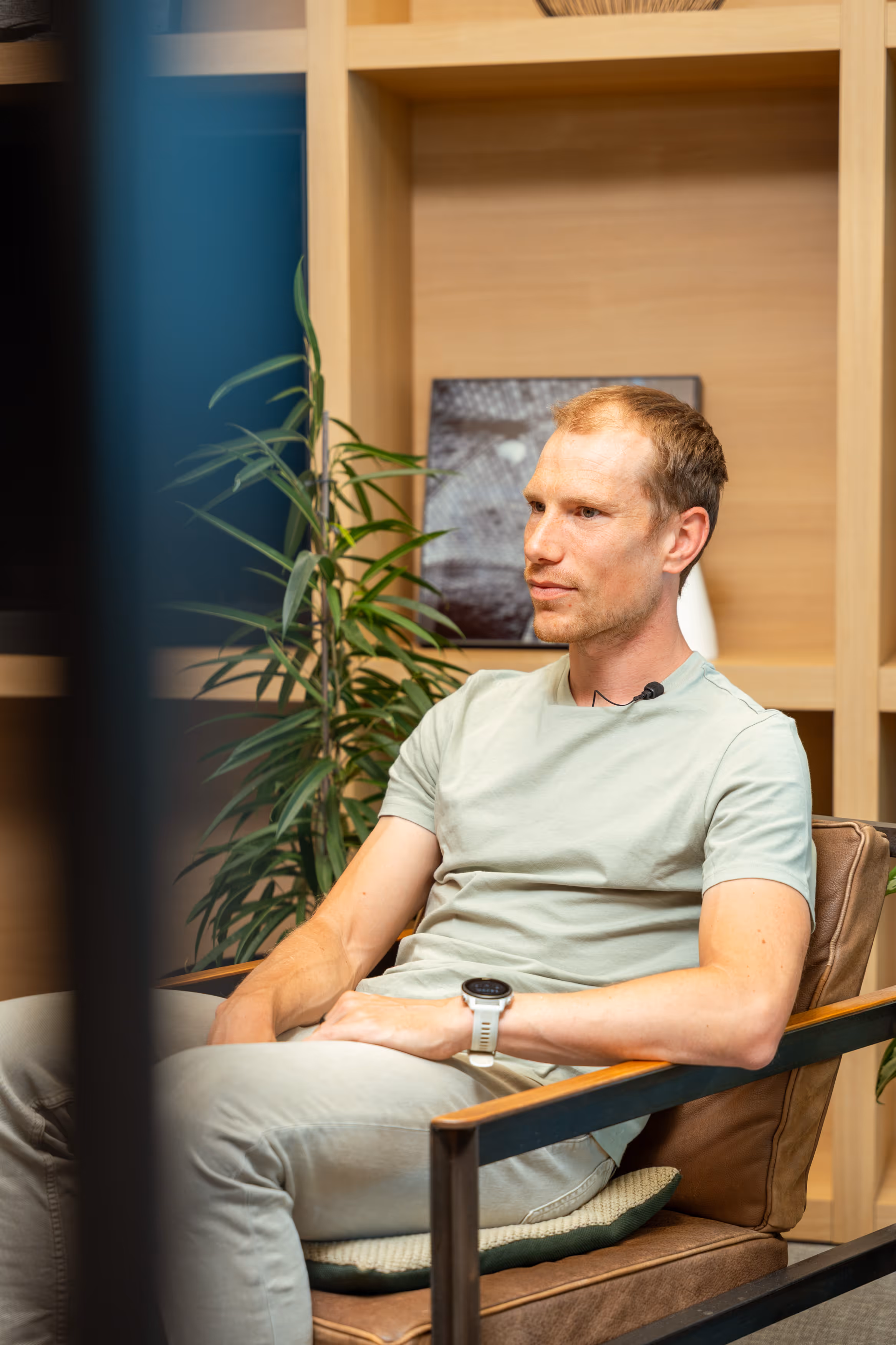 Man wearing a light green shirt and smartwatch sitting on a brown chair in a modern room.