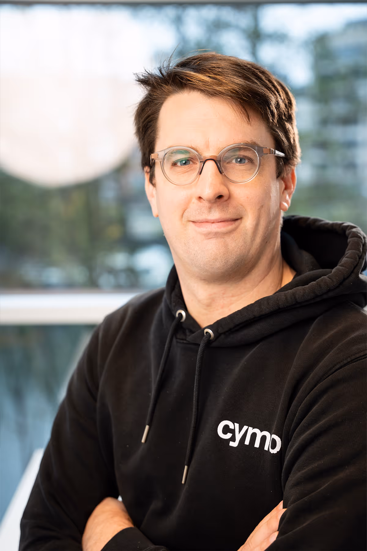 Man with glasses wearing a black hoodie with cymo logo posing indoors with arms crossed.