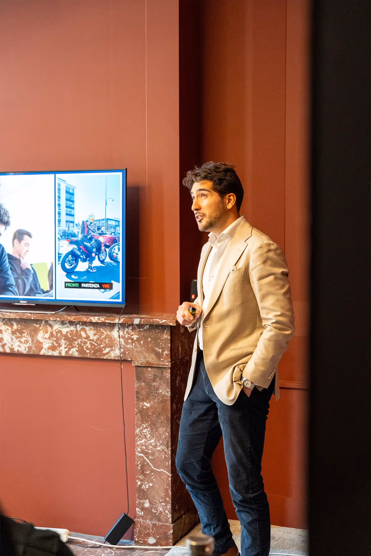 Man in beige blazer presenting next to a TV screen in a room with red walls and marble fireplace.