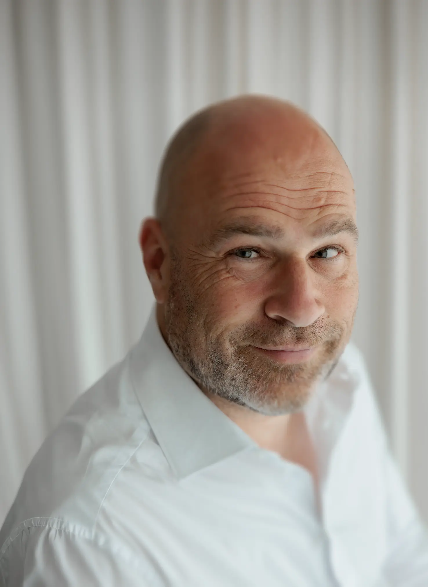 Smiling bald man with beard wearing a white shirt in front of white curtains.