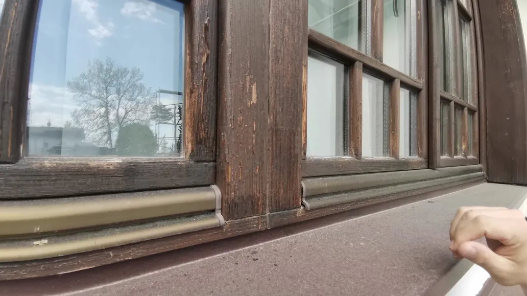 Staré dřevěné okno před renovací. Nátěr na rámu a křídlech je oloupaný, dřevo je zvětralé a popraskané. Pod křídly jsou kovové okapnice a plechový parapet.
