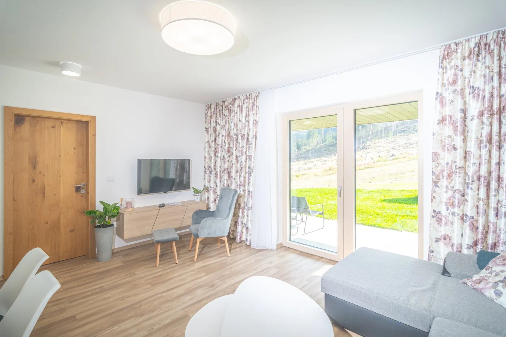 Obývací pokoj apartmánu s prosklenými balkonovými dveřmi v bílém rámu, za kterými je terasa se židlí a trávník. Vlevo dubové interiérové dveře, u stěny šedé křeslo a televizní stolek.