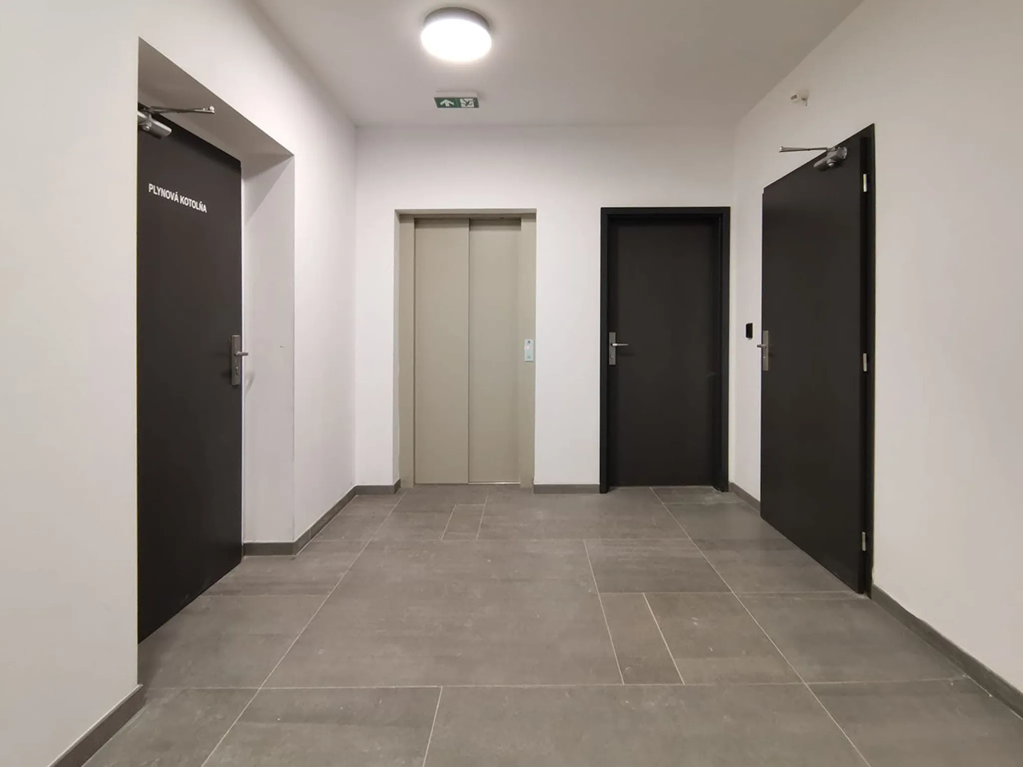 Chodba s dlaždicovou podlahou a černými dveřmi, dvoje z nich mají nahoře na křídle samozavírač. Dveře vlevo mají nápis označující plynovou kotelnu, uprostřed jsou dveře výtahu.