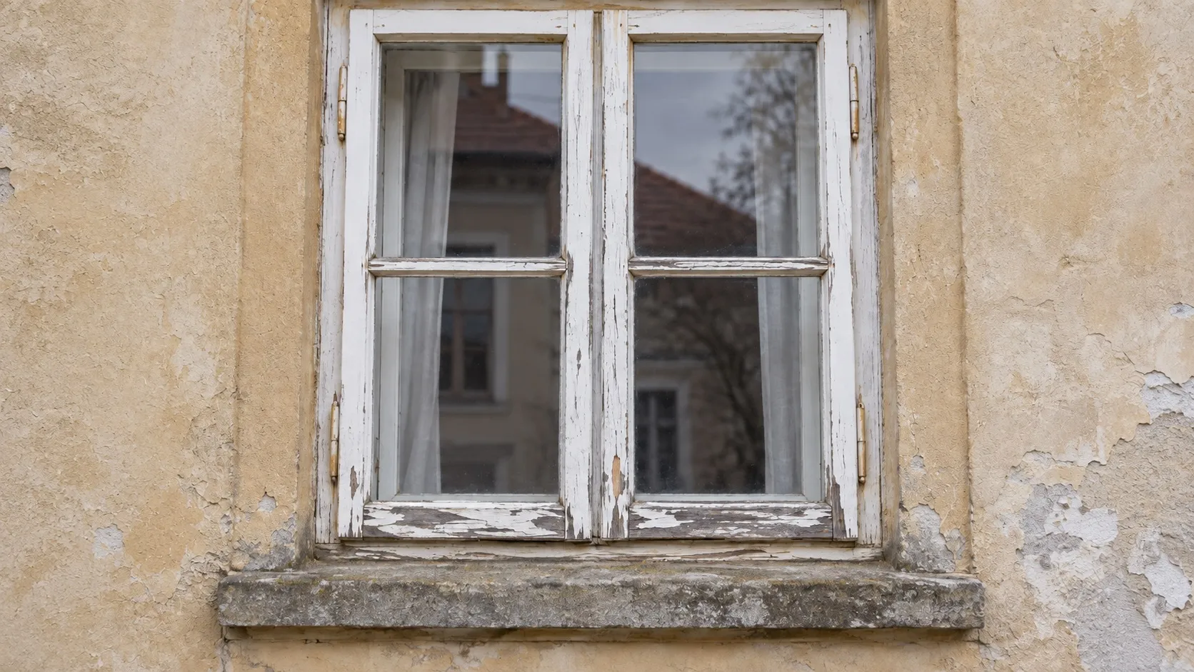 Staré dřevěné okno se dvěma křídly s příčkou na omítnuté fasádě. Bílý nátěr na spodních vlysech se odlupuje a prosvítá šedé dřevo, pod oknem je kamenný parapet.