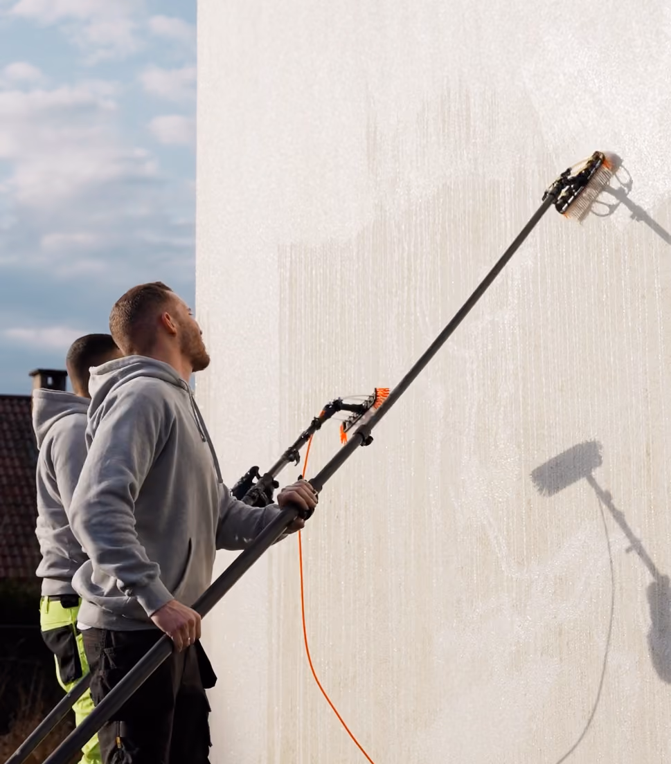 Twee mannen reinigen een witte buitenmuur met telescopische borstels terwijl de zon schijnt en een blauwe lucht zichtbaar is.
