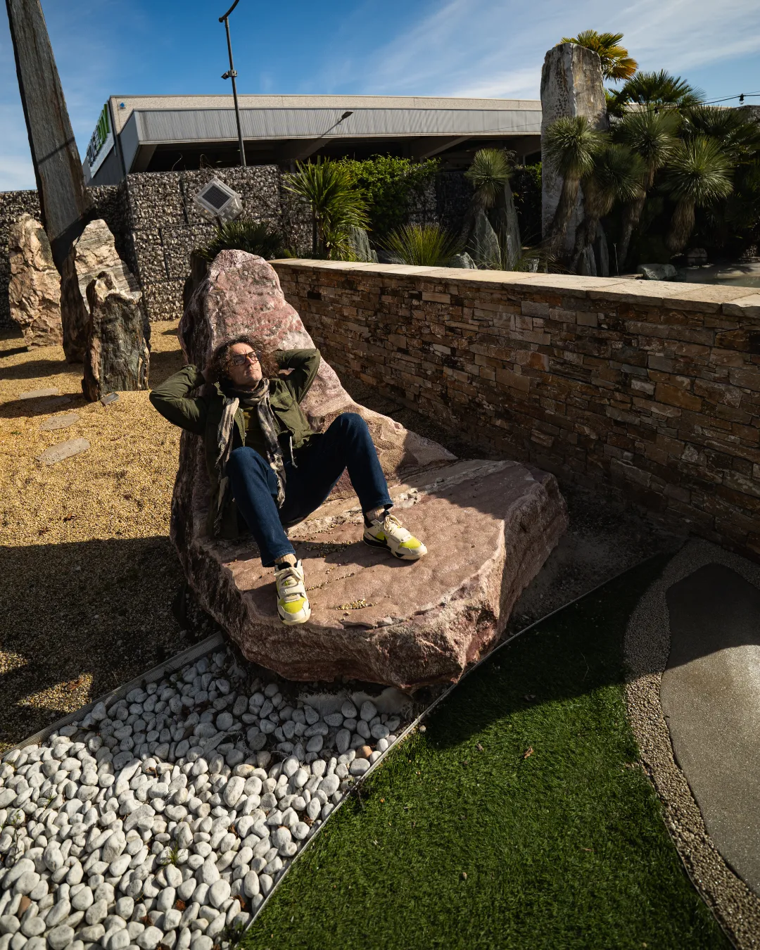 Una persona rilassata seduta su una grande pietra modellata a forma di sedia in un giardino soleggiato.