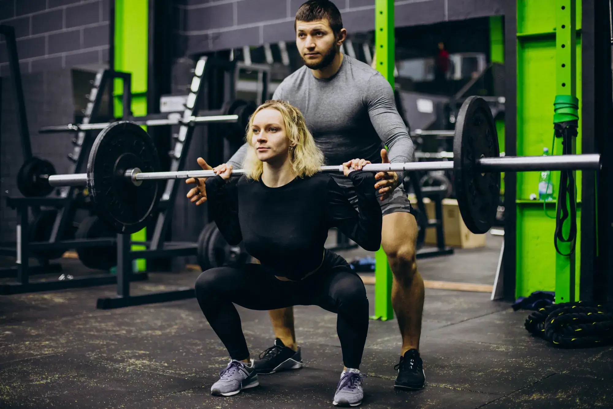 Woman squats with a barbell on her shoulders while a man spotter supports her in a gym.
