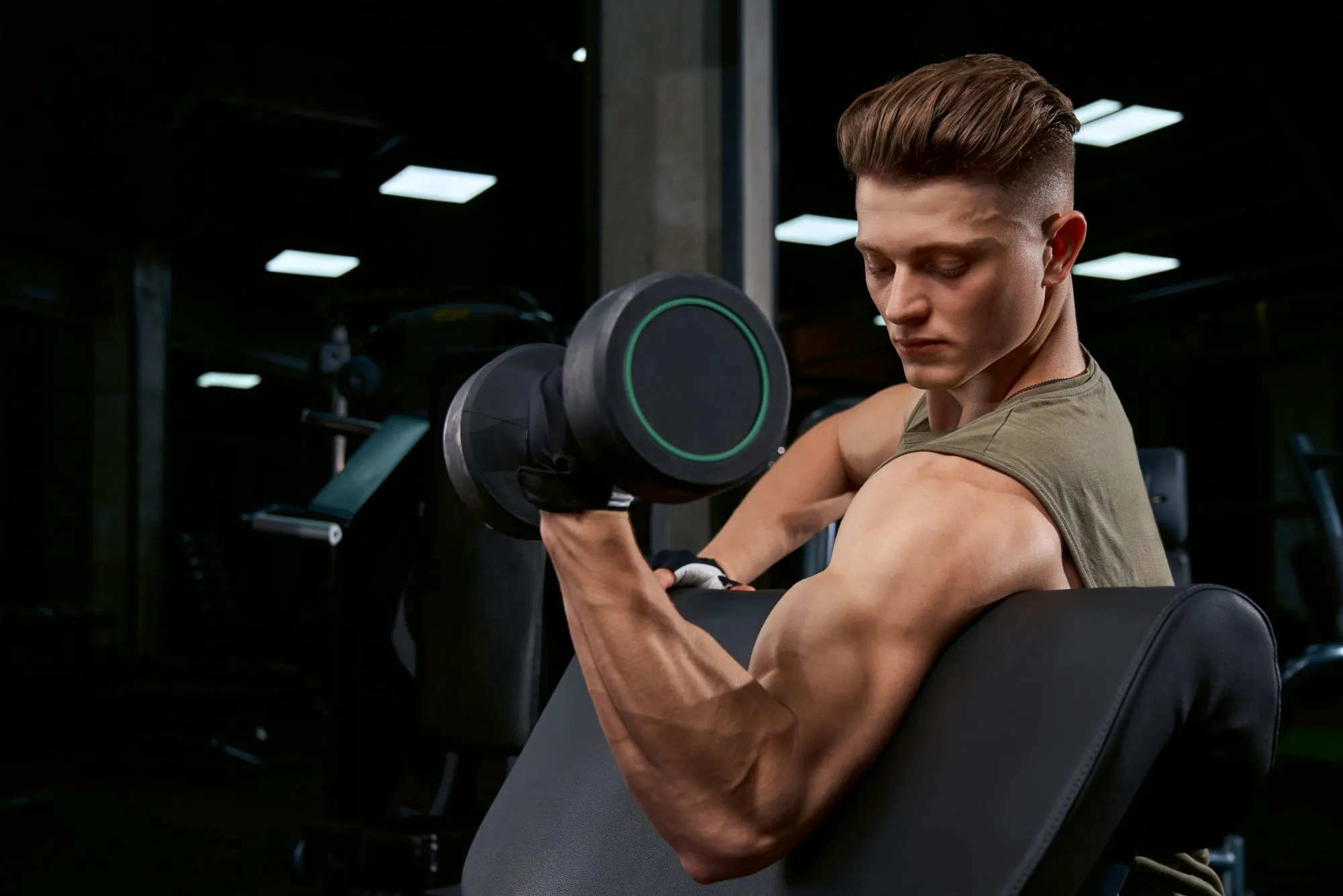 Young man in green tank top doing bicep curls with dumbbell on preacher curl bench in gym.