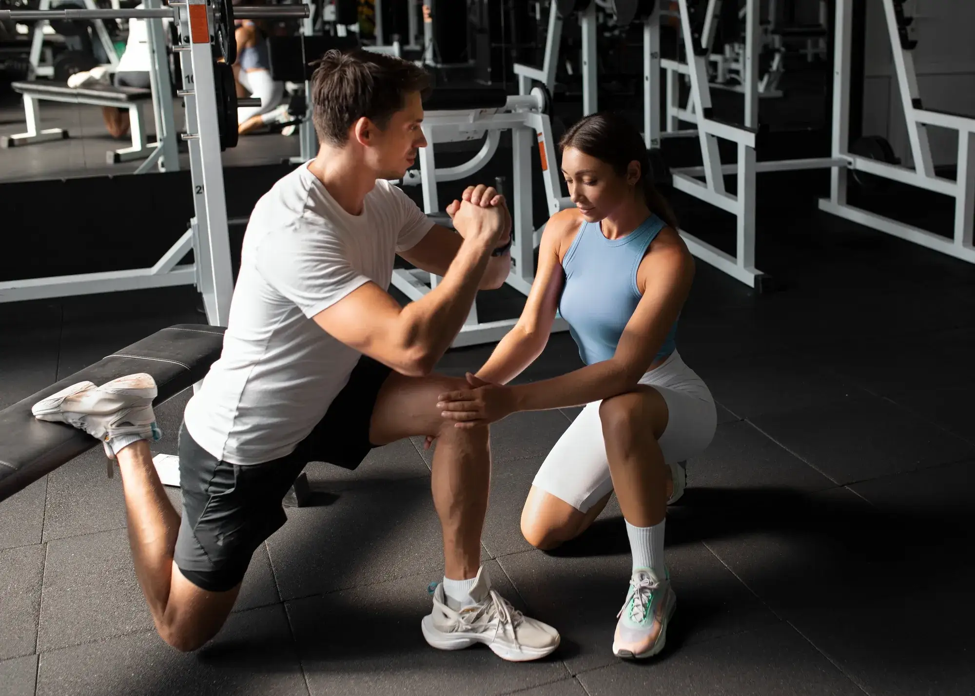 Man in white shirt kneeling with one leg while woman supports his knee in gym setting.
