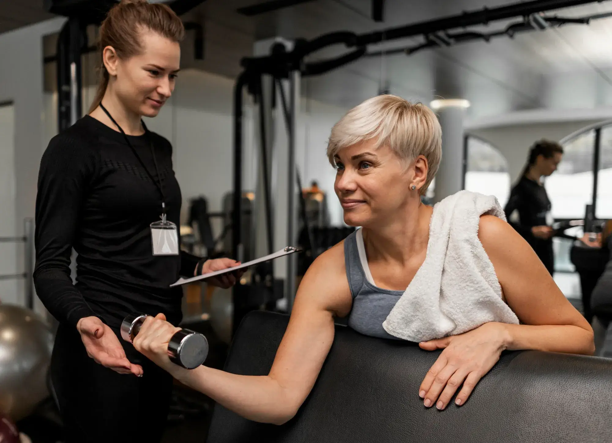 Woman lifting dumbbell with trainer guiding her in a gym, a towel draped over her shoulder.