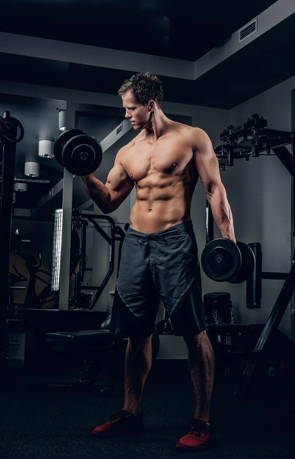 Muscular shirtless man lifting dumbbells in a dimly lit gym with workout equipment around him.