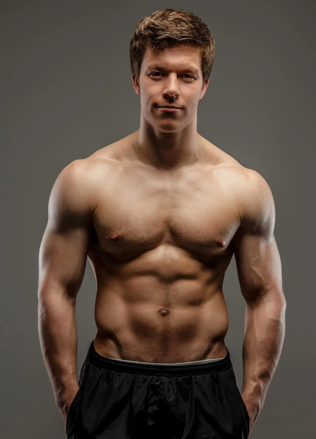 Muscular shirtless man with defined abs wearing black shorts standing against gray background.