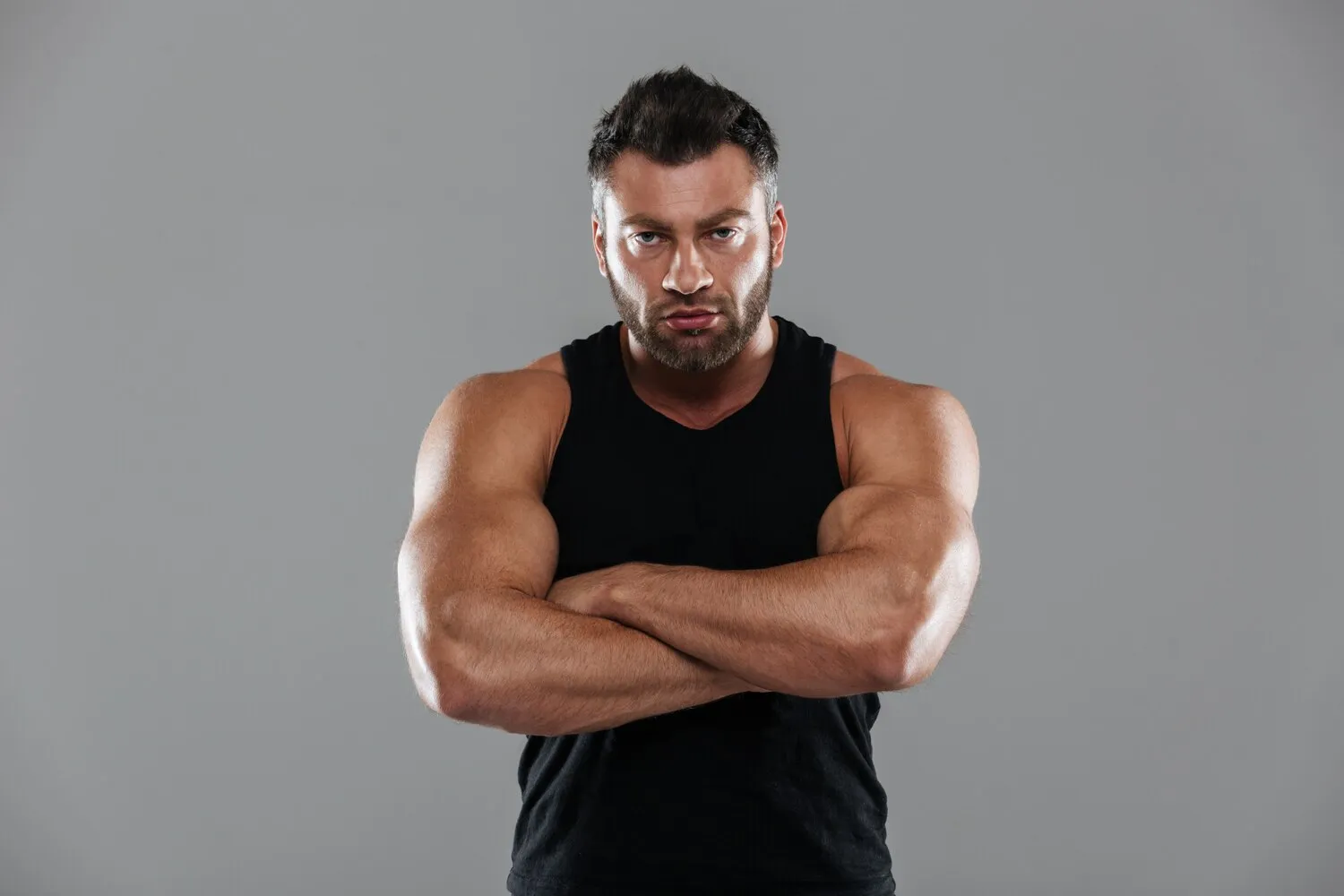 Muscular man in a black sleeveless shirt with arms crossed against a gray background.