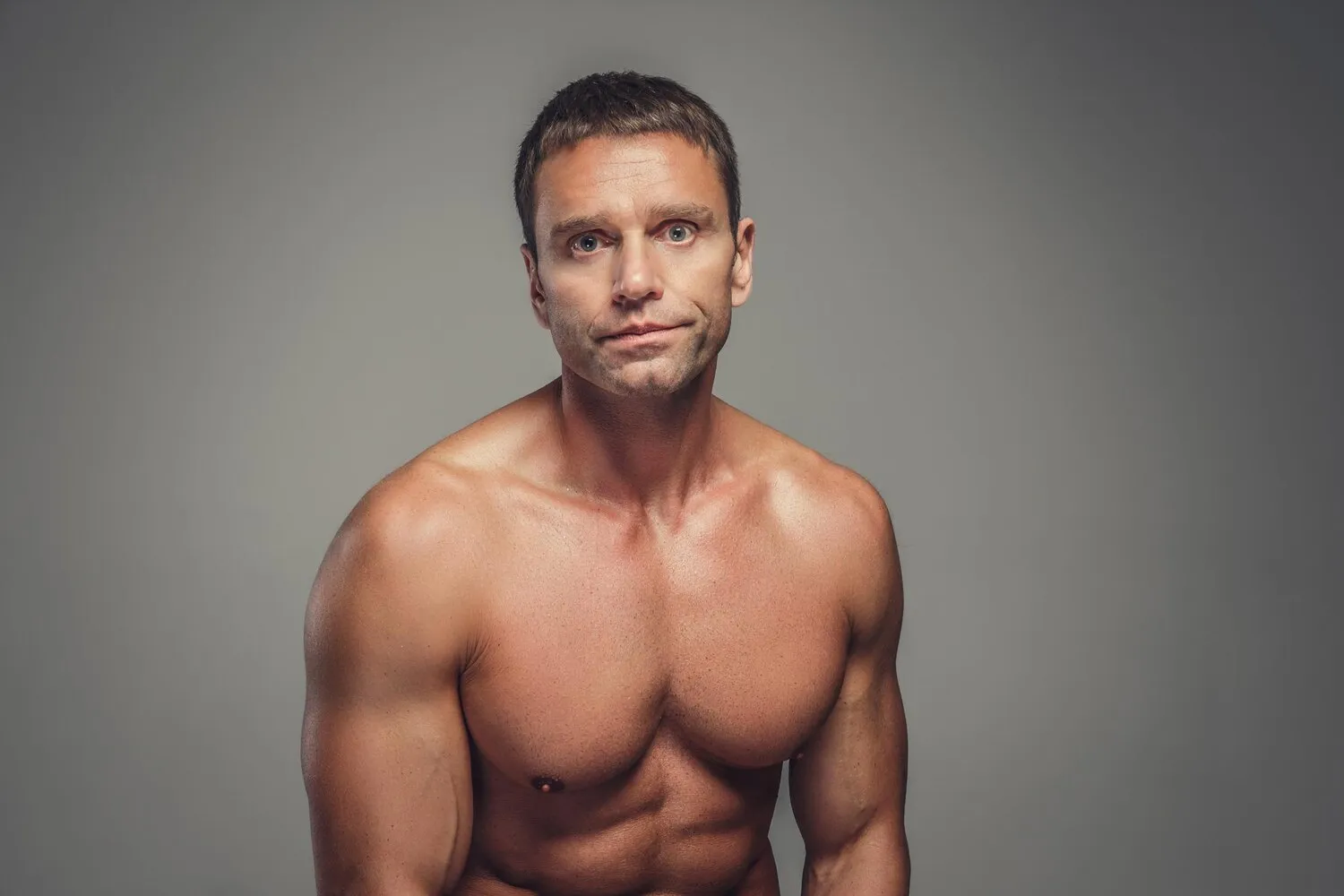 Shirtless muscular man with short hair looking forward against a plain gray background.
