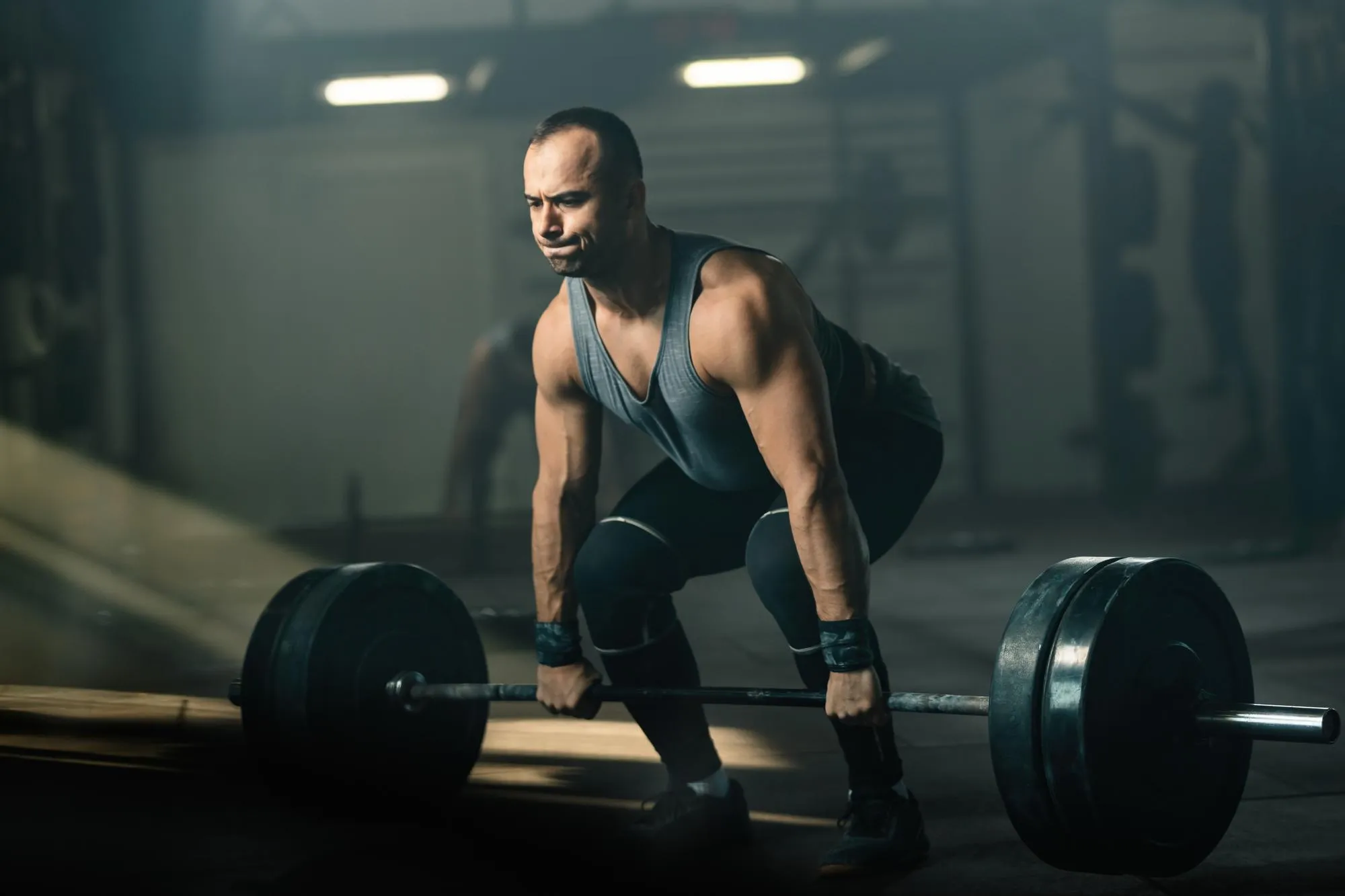 Muscular man in gym lifting heavy barbell with focused expression in dimly lit environment.