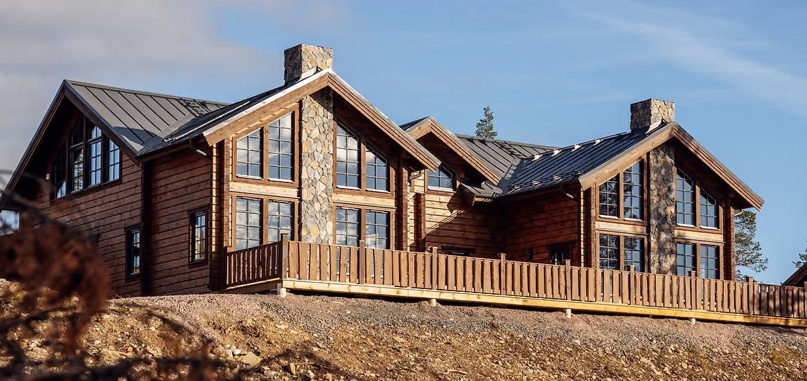 Large wooden cabin with stone chimneys and many windows under a clear blue sky.