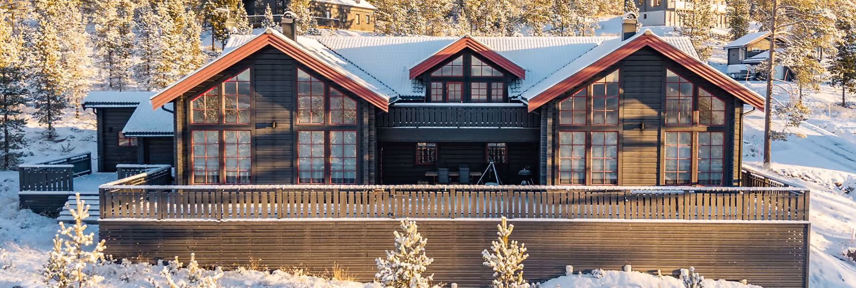 Large wooden cabin with snowy roof and trees in a winter forest setting at sunset.