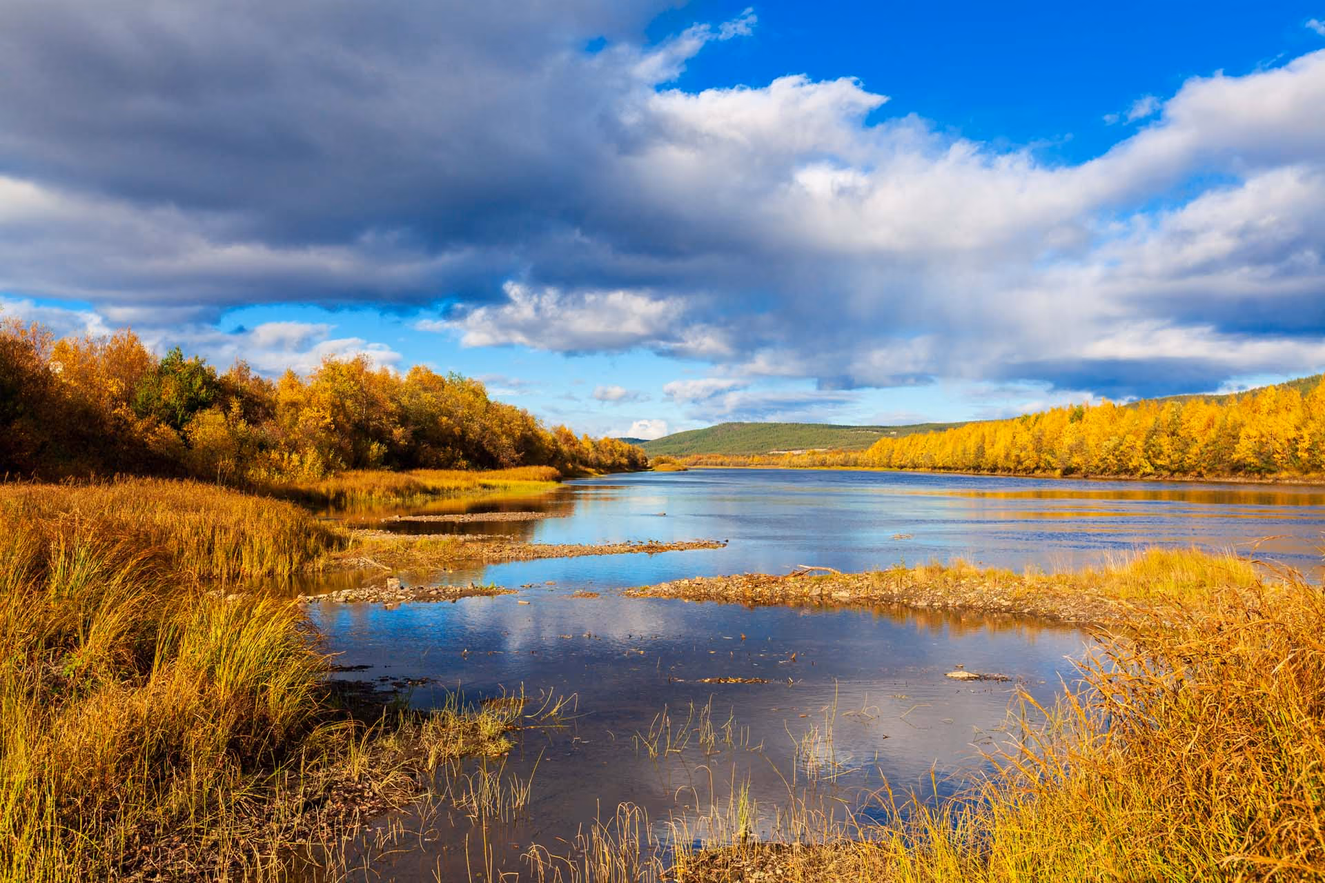 Calm river reflecting clouds, surrounded by autumn trees and grass under a partly cloudy sky.