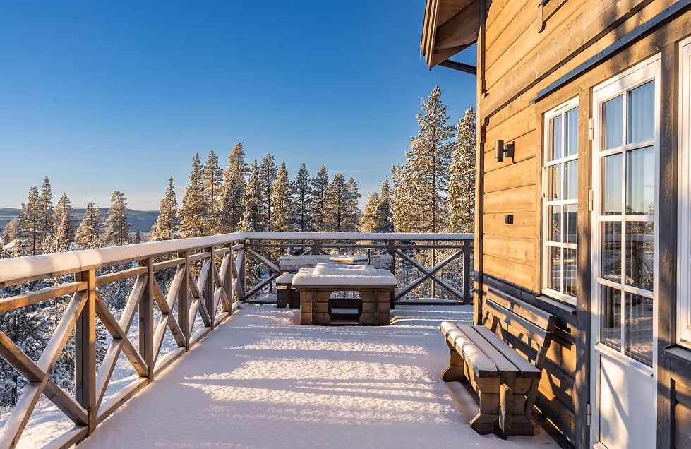 Snow-covered wooden deck with bench and table outside a cabin surrounded by pine trees under blue sky.